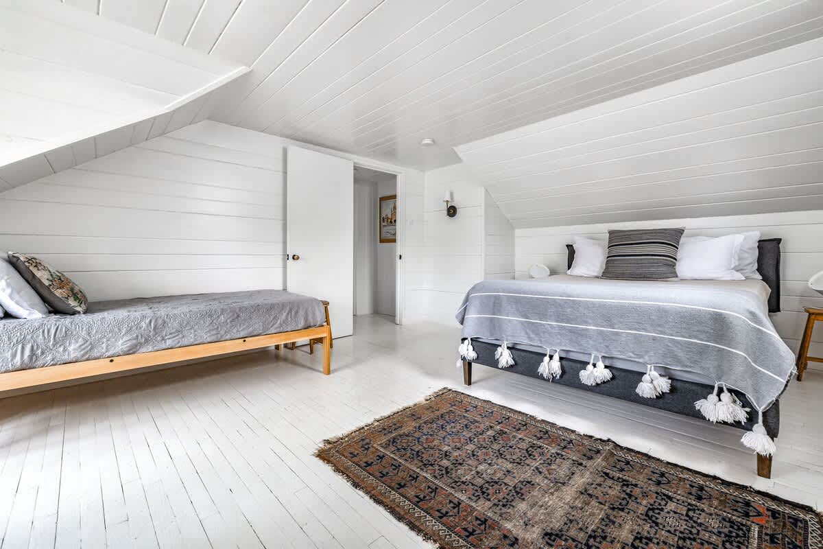 Bright attic bedroom with white shiplap walls and painted floorboards, featuring a queen-sized bed with a grey blanket and decorative tassels, and a single bed (Bedroom 1) in the background. A patterned rug lies on the floor.