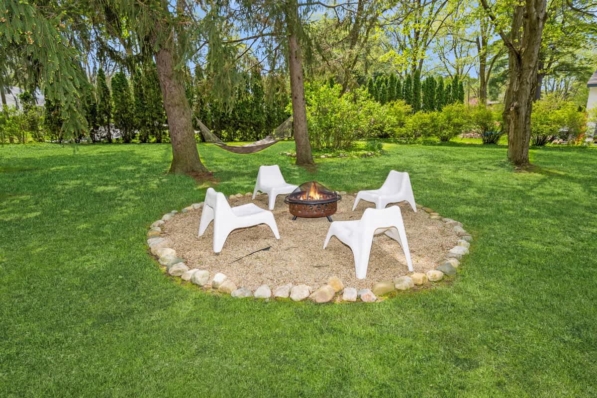 Outdoor fire pit area with a round metal fire bowl on a gravel base, encircled by small rocks, and four modern white chairs arranged around it on a vibrant green lawn.
