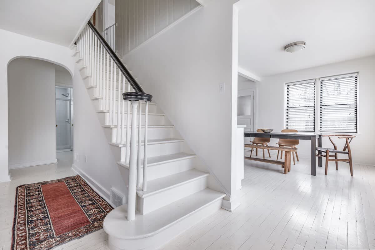 Bright foyer area featuring a white staircase with a black handrail and a decorative newel post, leading upstairs. A patterned area rug is placed on the white wood floor, and an arched doorway is visible to the left.