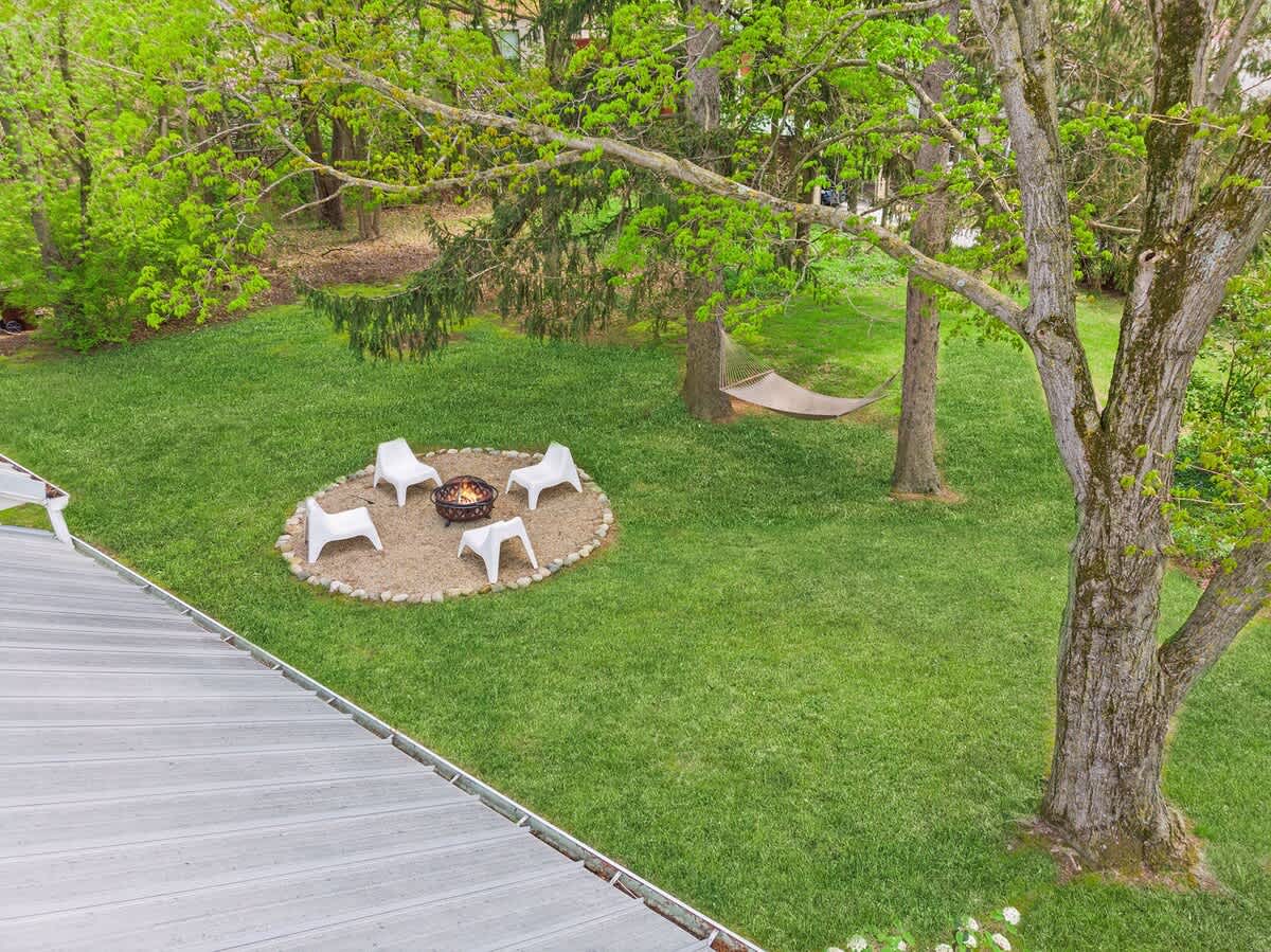 Elevated view of the expansive green backyard, featuring a circular fire pit area with white chairs, a hammock strung between trees, and mature trees providing shade.