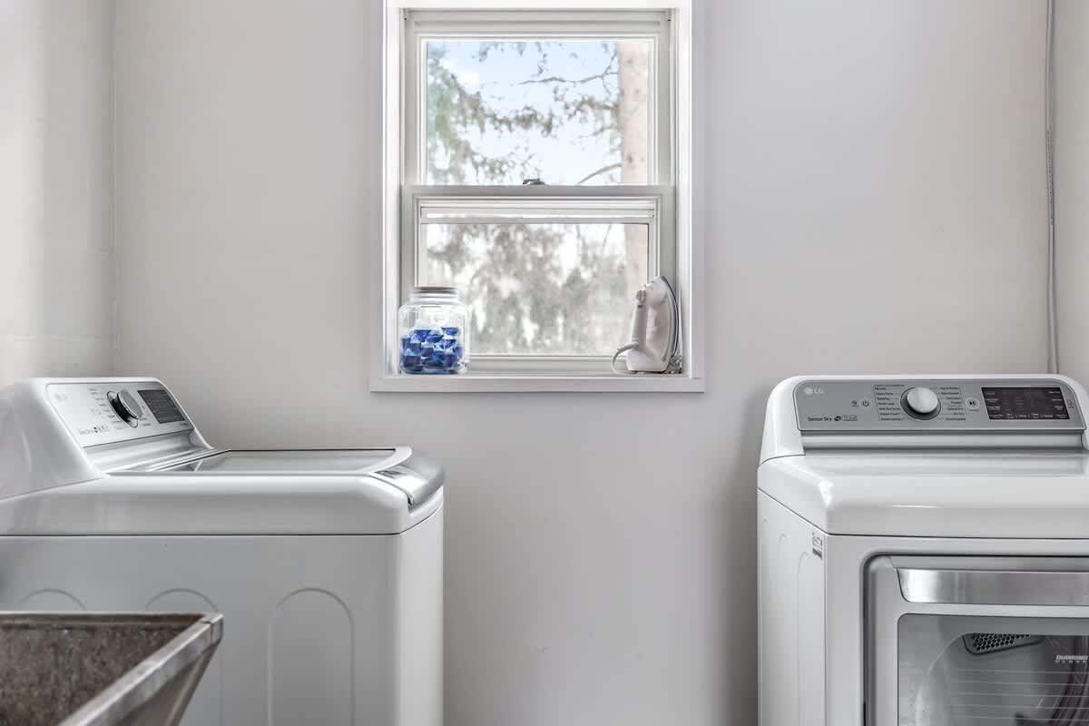 Functional laundry room with a white top-load washing machine and a front-load dryer, positioned beneath a window. A jar of blue laundry pods sits on the windowsill.