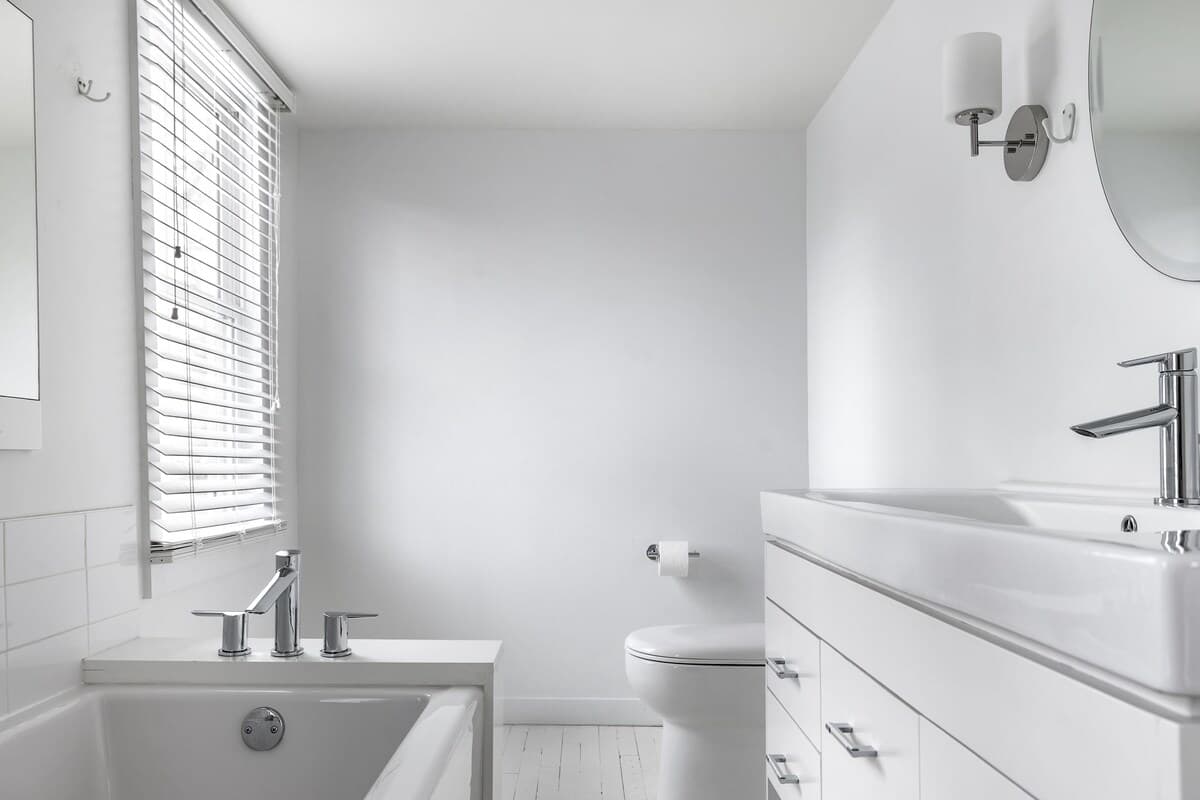 Modern white bathroom with a deep soaking tub, a toilet, and a vanity with a rectangular sink and chrome faucet. A window with blinds provides natural light.