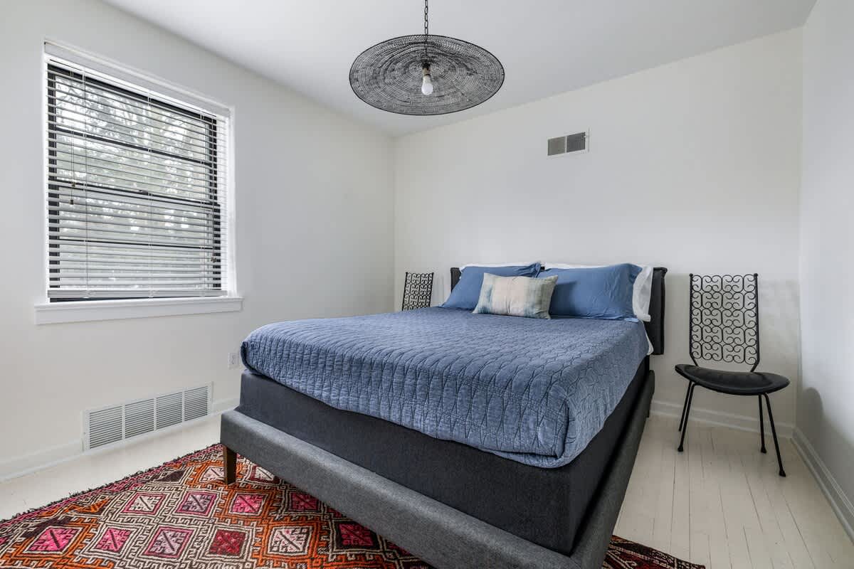 Bedroom with a queen-sized bed featuring a blue quilted comforter and decorative pillows, a colorful patterned rug, and a window with blinds, all set on light hardwood floors.
