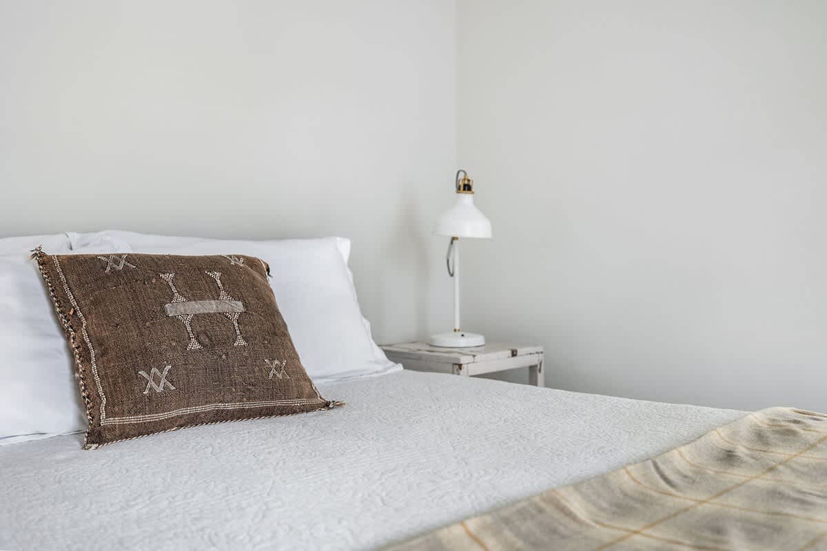 A close-up view of the bed in a bright bedroom, featuring crisp white bedding, a textured brown throw pillow with a geometric pattern, and a white table lamp on a distressed white nightstand.