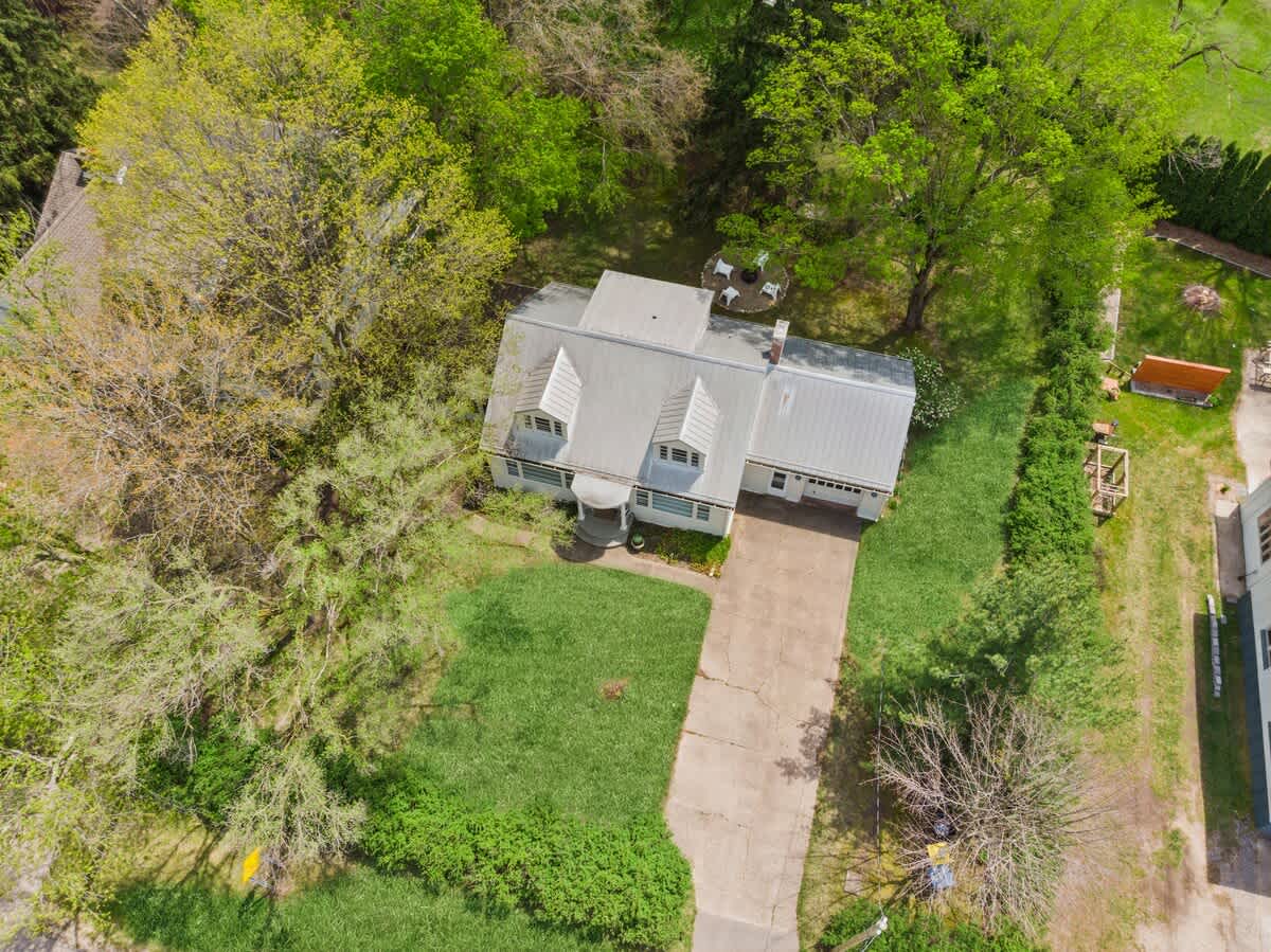 Aerial view showcasing a charming white house with a light-colored metal roof and dormer windows, a concrete driveway, and extensive green lawn and mature trees surrounding the property.