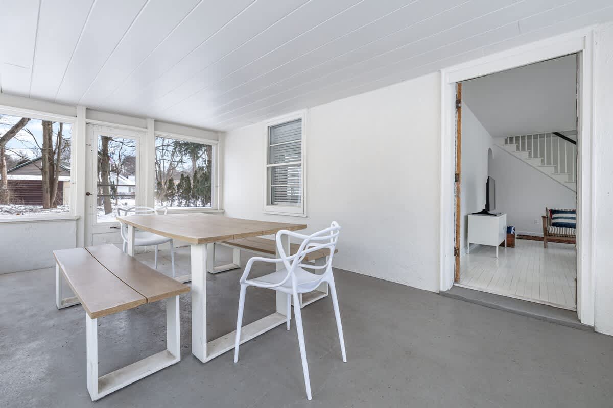 A bright enclosed porch or sunroom with white paneled ceilings and walls, featuring a light wood dining table with two matching benches and two modern white chairs, surrounded by large windows.