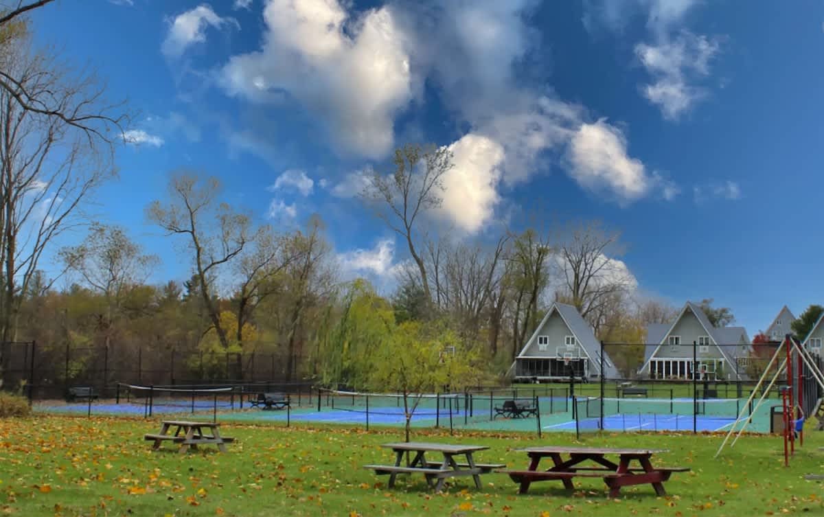 Two outdoor tennis courts with blue and green surfaces, enclosed by a fence, with picnic tables on a grassy area in the foreground and A-frame chalets visible through the trees in the background.