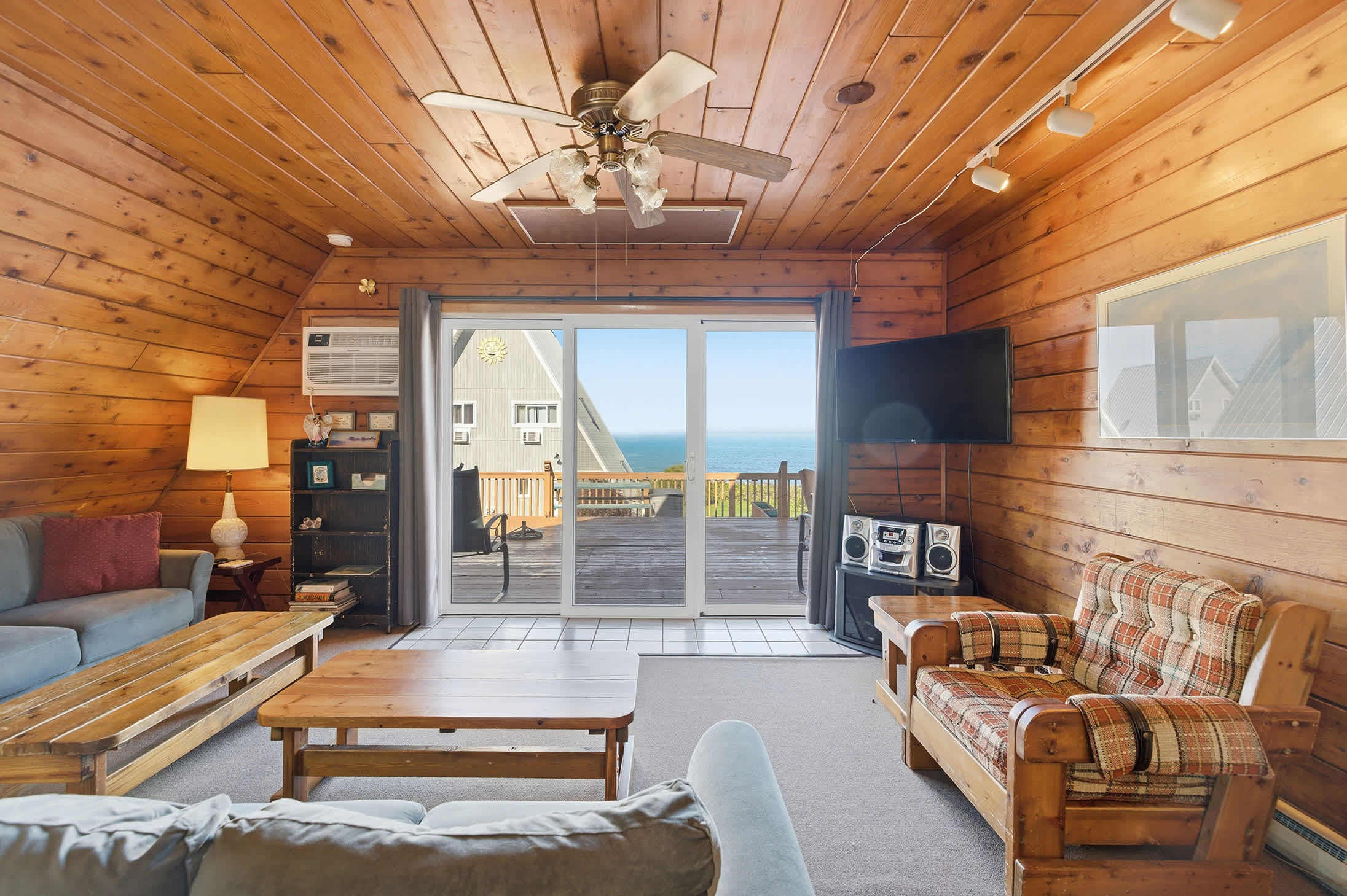 Wide shot of the living room with rustic wood-paneled walls and ceiling, featuring two blue sofas, a plaid armchair, two wooden coffee tables, and a sliding glass door opening to a deck with a partial view of another building and the lake.