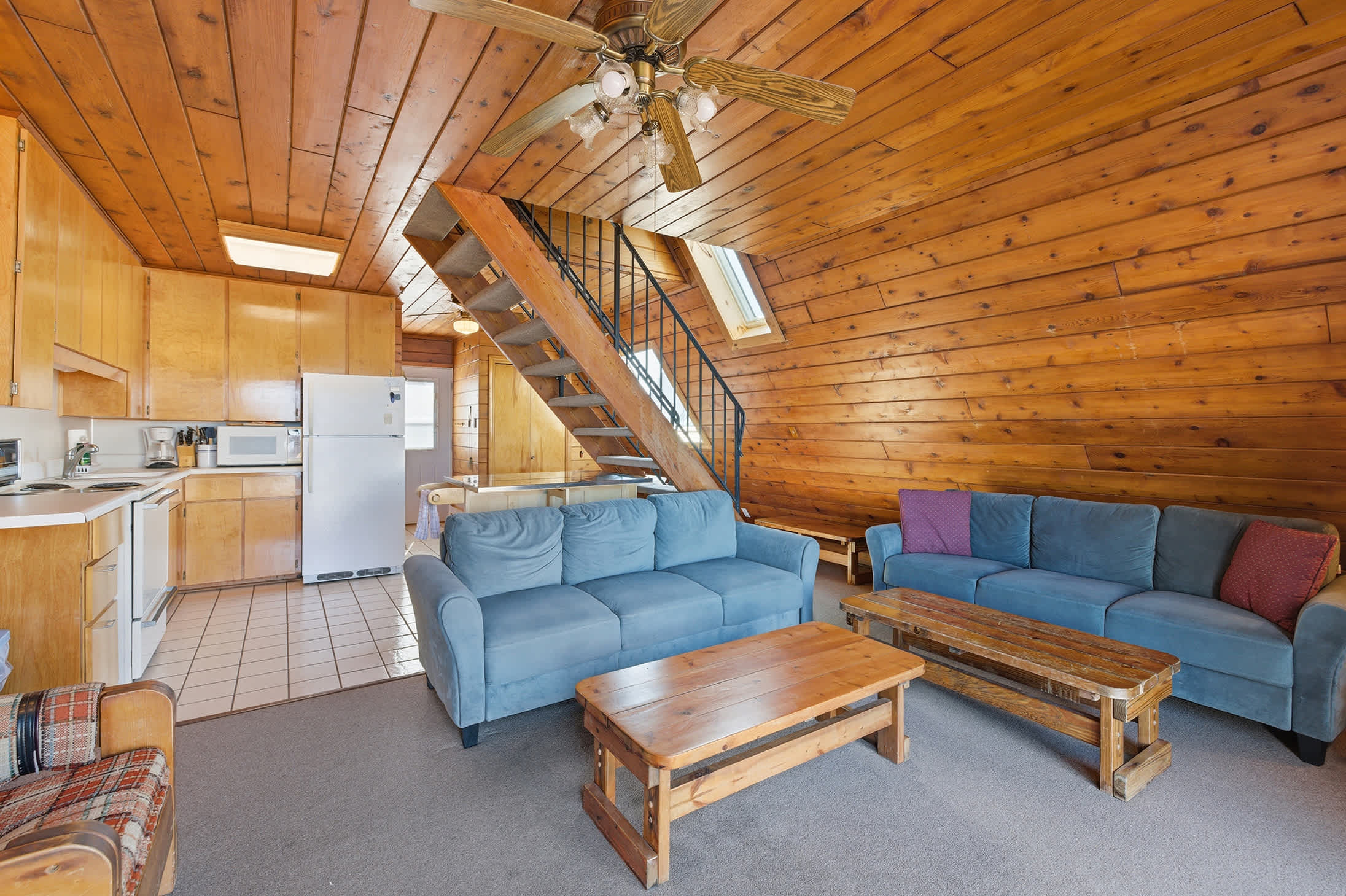 View of the living room from a different angle, showcasing two blue sofas, a wooden coffee table, and a black metal staircase leading upstairs. The room has consistent wood-paneled walls and ceiling, with a kitchen area visible in the background.