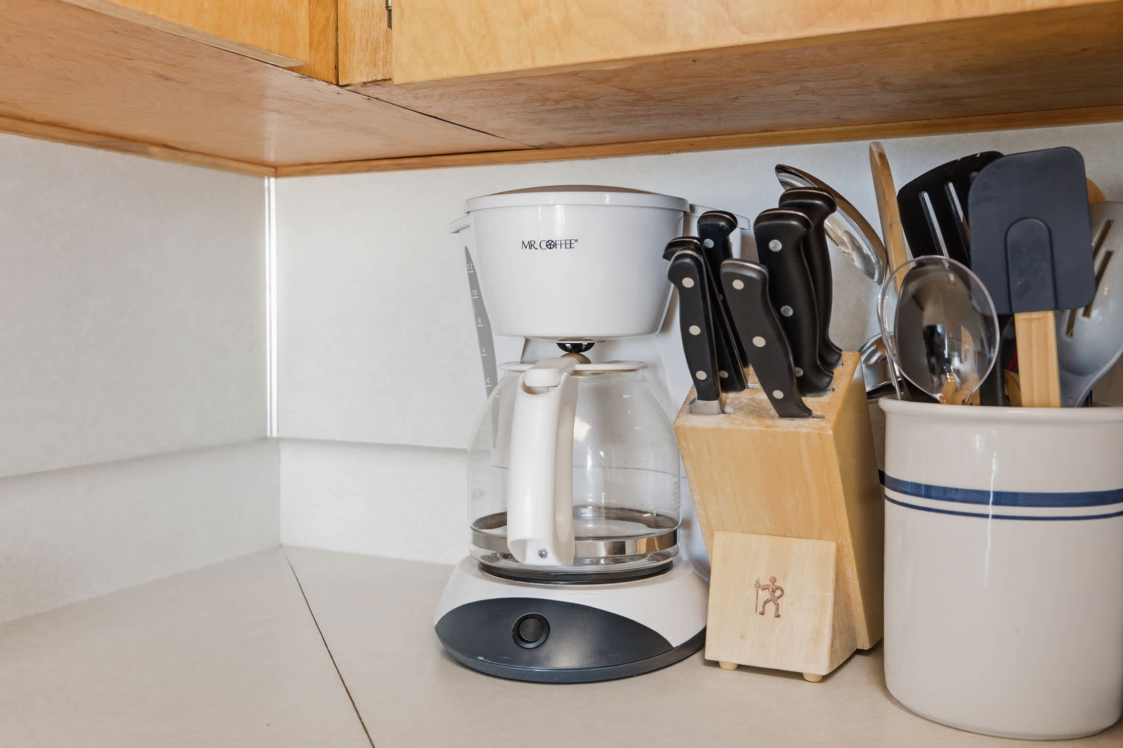 Close-up of a kitchen counter showing a white Mr. Coffee coffee maker, a wooden knife block with various knives, and a ceramic utensil holder filled with cooking tools.