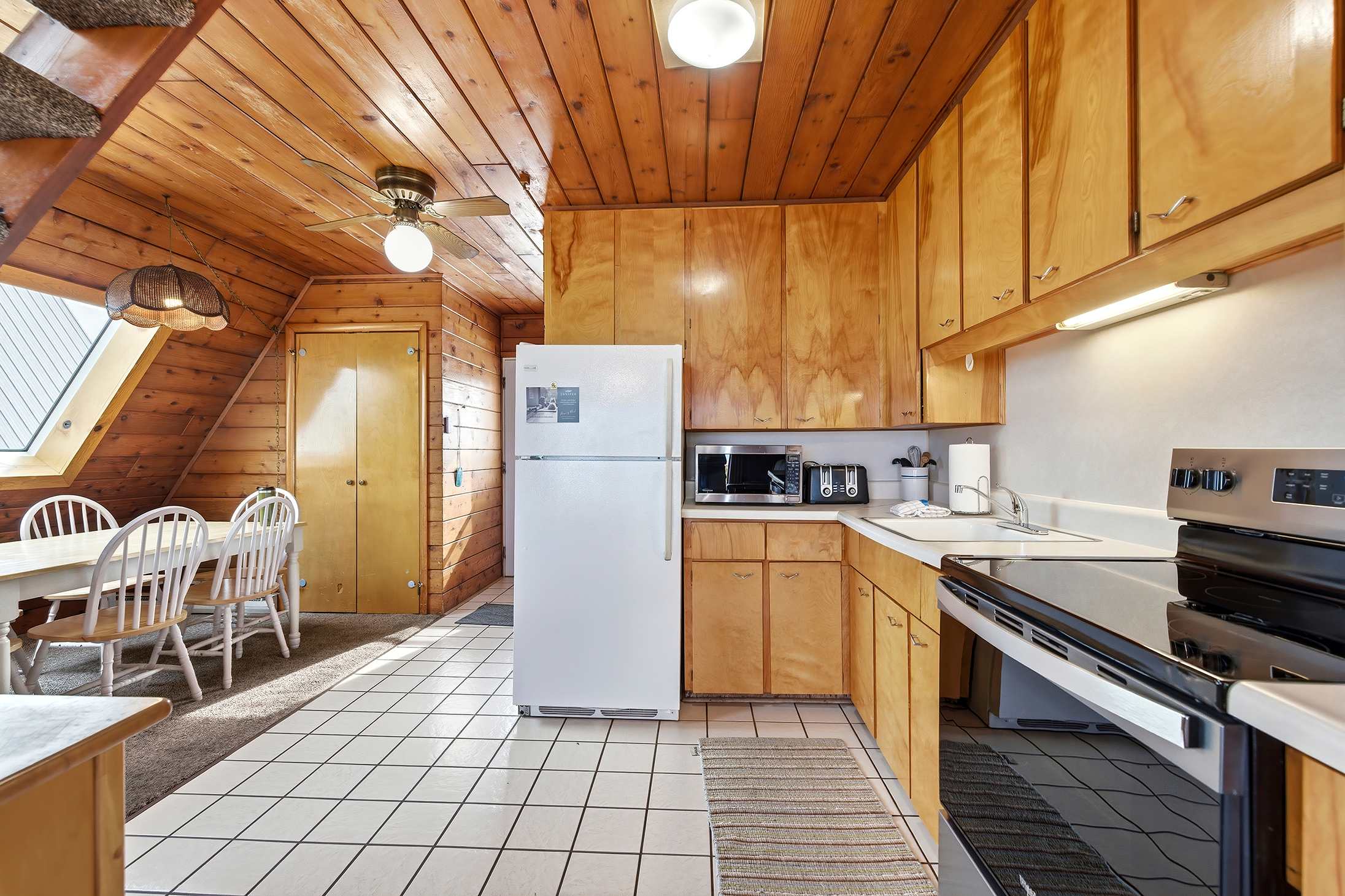 View of the kitchen and adjacent dining area, featuring light wood cabinetry, white appliances, and white tiled floors in the kitchen. The dining area has a light wood table and white chairs, with knotty pine walls and ceiling throughout.