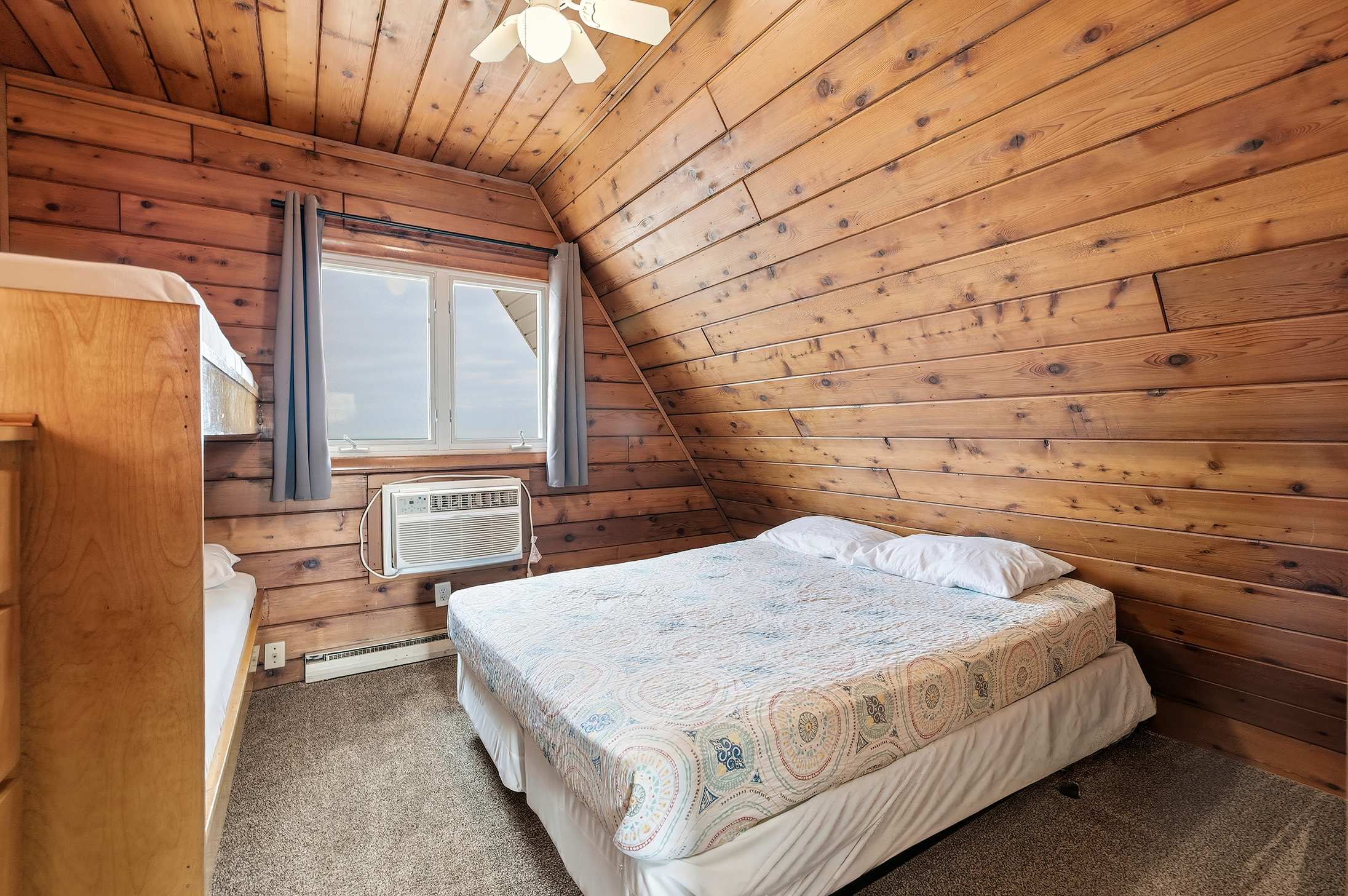 Cozy bedroom featuring rustic wood-paneled walls and a sloped ceiling, a queen bed with a patterned quilt, and a window with an AC unit. A bunk bed is partially visible on the left.