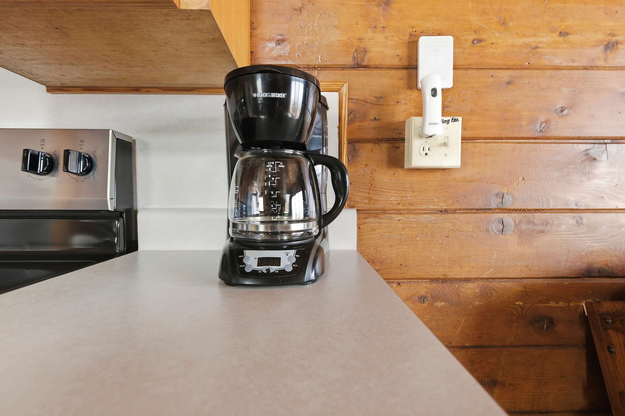 Close-up of a black coffee maker on a white countertop in a kitchen with knotty pine walls. A wall outlet with a nightlight is visible next to it.