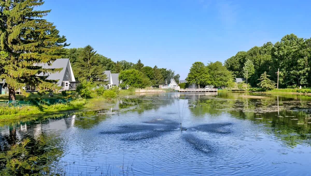 Picturesque view of a tranquil pond with a decorative fountain, bordered by lush green trees and several A-frame chalets under a clear blue sky.
