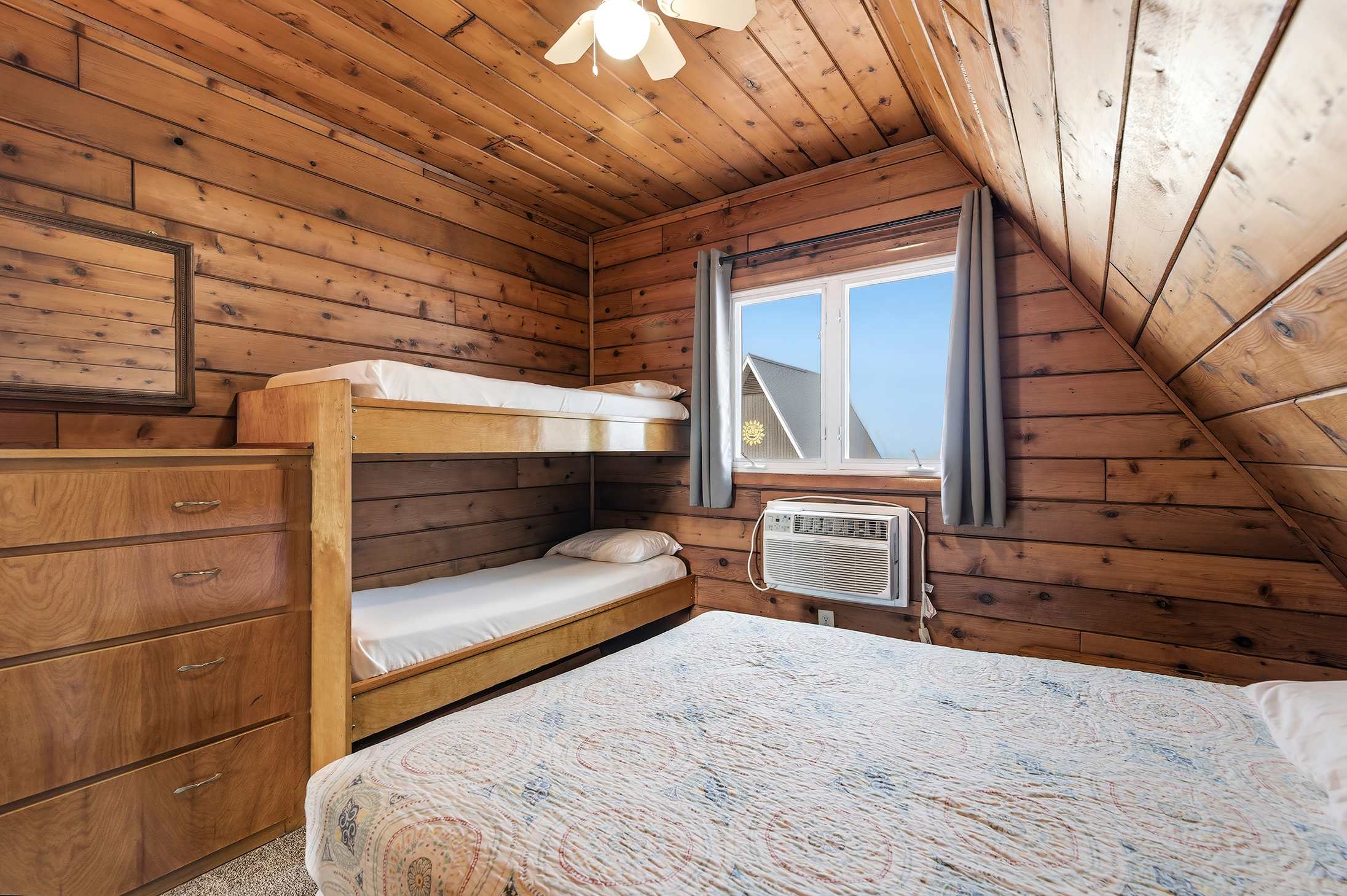 Cozy bedroom with knotty pine walls and a sloped ceiling, featuring a queen-sized bed with a blue and white patterned quilt, and a wooden bunk bed. A window offers an outdoor view, and an air conditioning unit is installed below it.