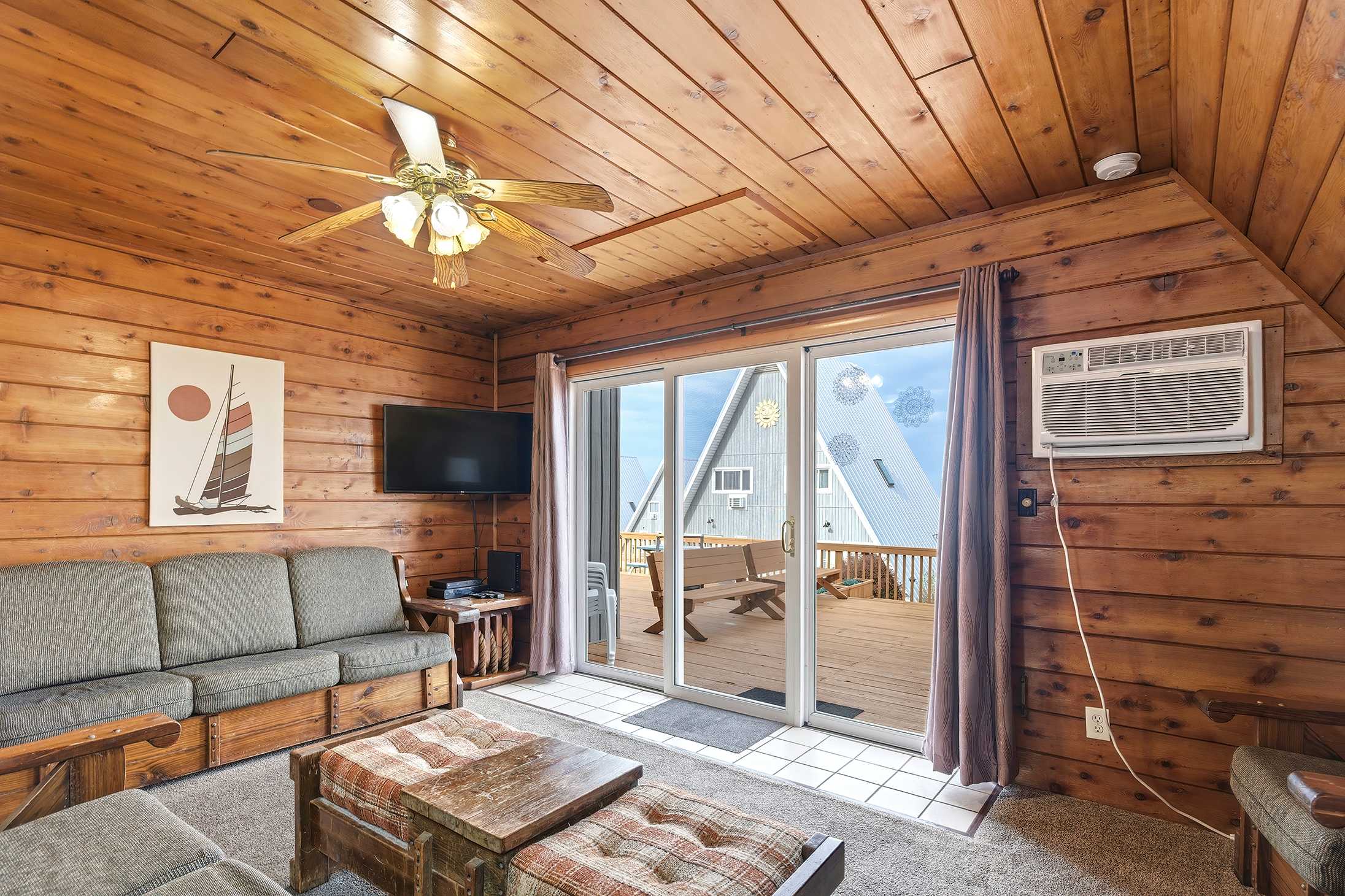 Living room with knotty pine walls and ceiling, featuring a gray sofa and a wooden coffee table with plaid ottomans. Sliding glass doors open to a deck with views of another A-frame chalet, and a wall-mounted TV is visible.