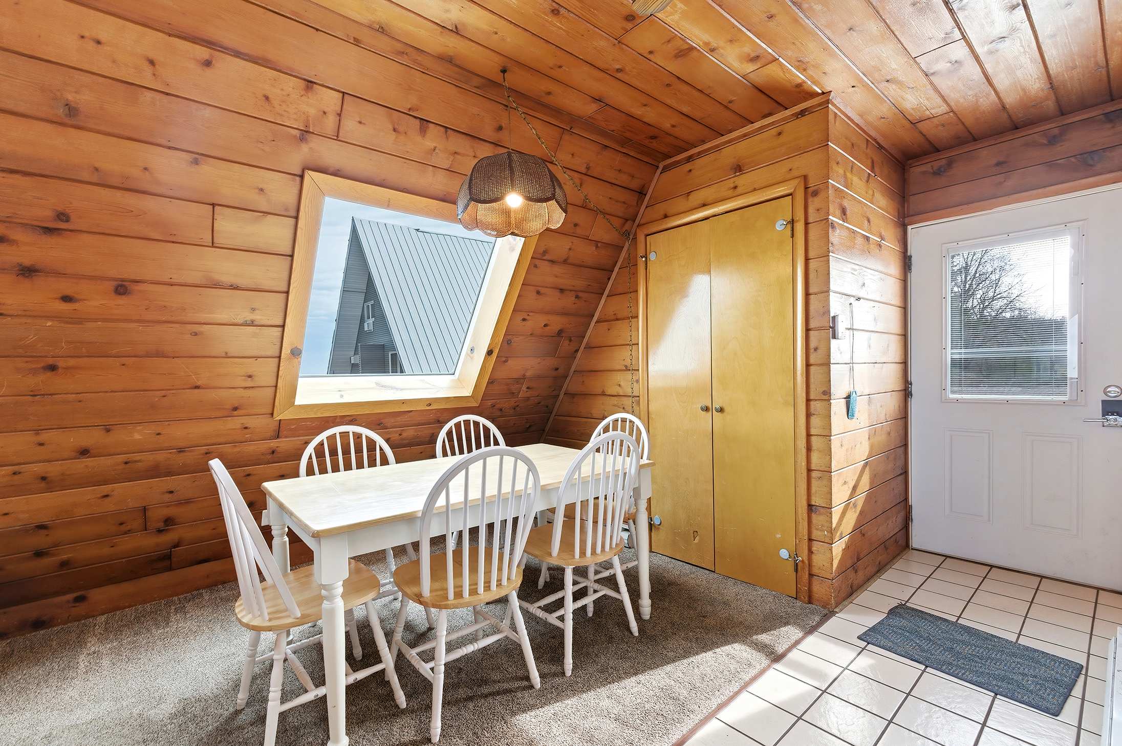 Dining area with knotty pine walls and a sloped ceiling, featuring a light wood dining table with four white chairs. A large angled window provides natural light, and a white exterior door is visible.
