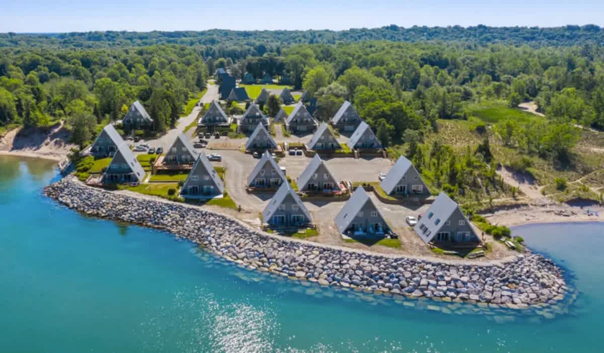 A wide aerial shot revealing a community of numerous A-frame houses arranged in rows along a picturesque blue-green lakefront. The property is bordered by a lush green forest, highlighting its secluded natural setting.