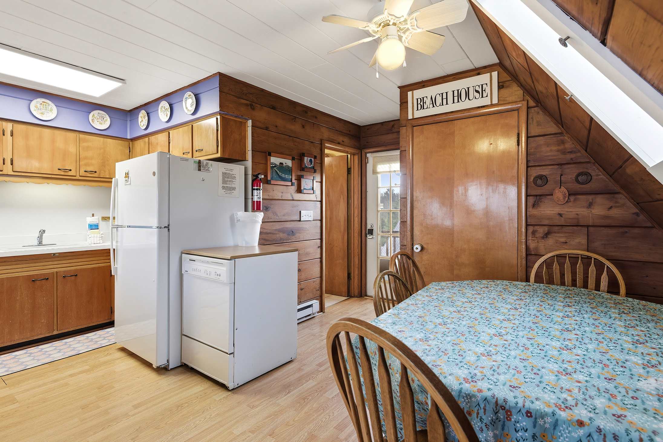 The kitchen area features light wooden cabinets and white appliances, with a purple accent wall above the cabinets. Adjacent is a dining table with a floral tablecloth and wooden chairs, all set against knotty pine walls.