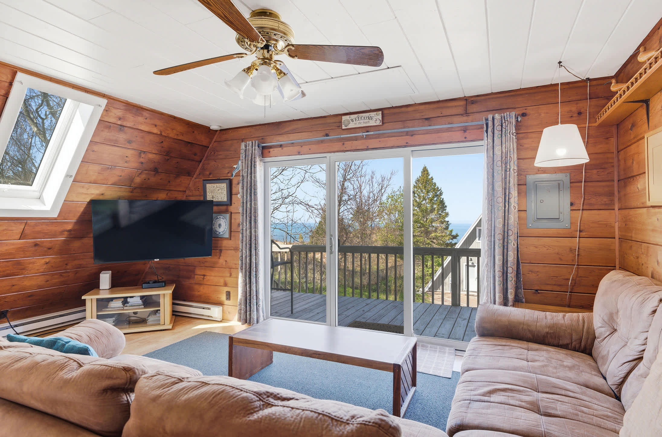 The living room features knotty pine walls and a white ceiling with a ceiling fan, centered around a flat-screen TV on a wooden stand. A large sectional sofa faces sliding glass doors that lead to a deck with views of trees and water.