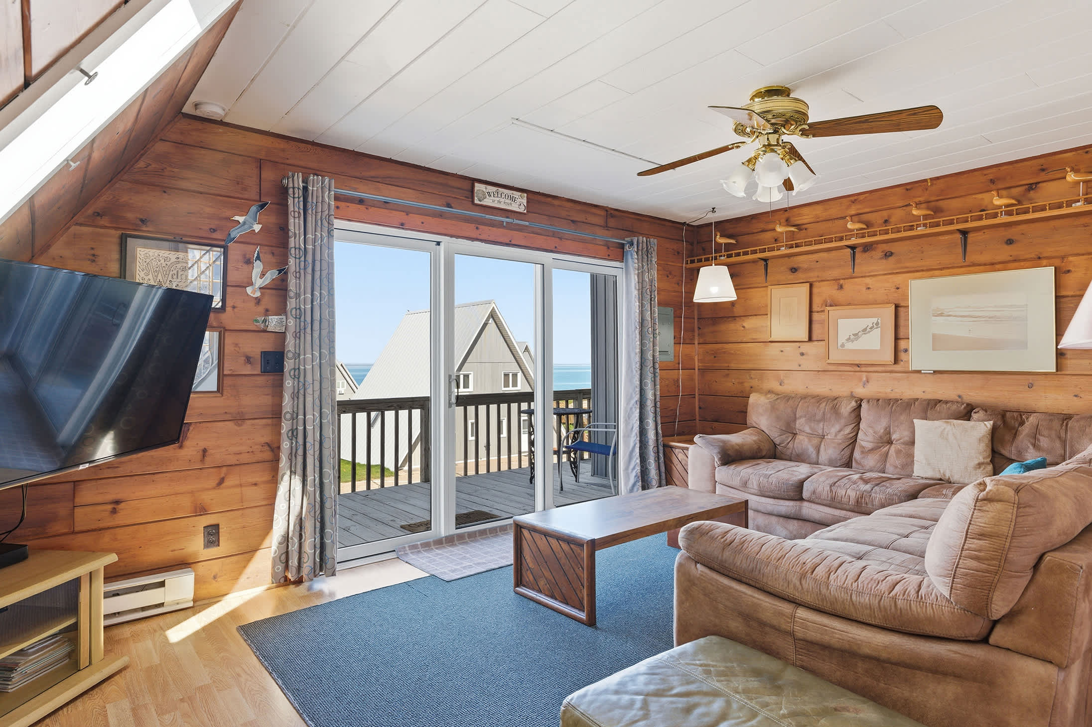 The living room showcases knotty pine walls and a white ceiling with a brass ceiling fan, featuring a large sectional sofa on a blue rug. Sliding glass doors provide access to a deck with views of other chalets and the water.