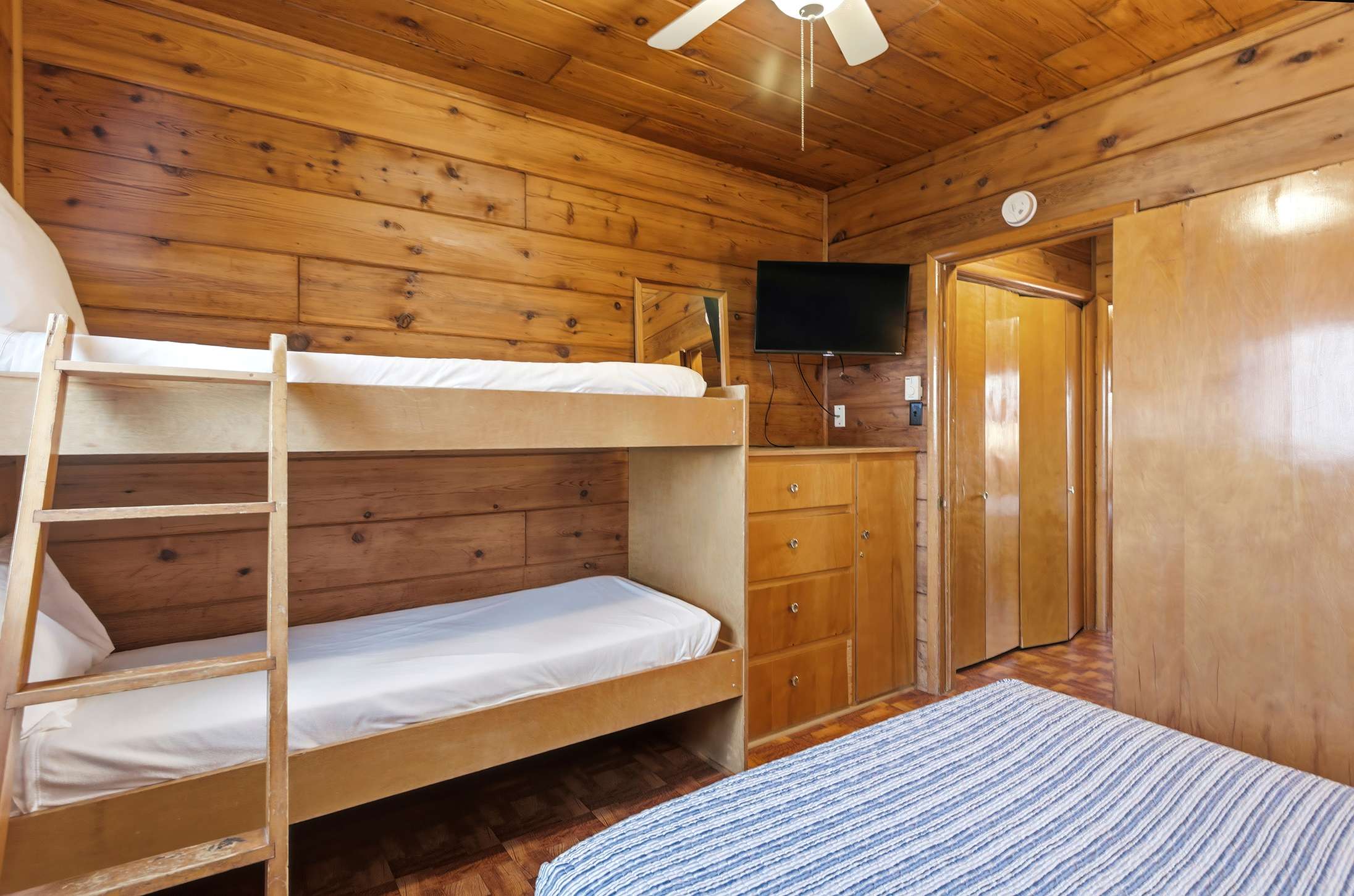 Bedroom-1 showcases knotty pine walls and a sloped ceiling, featuring a wooden bunk bed and a double bed. A small flat-screen TV is mounted on the wall above a dresser, and a closet door is visible.