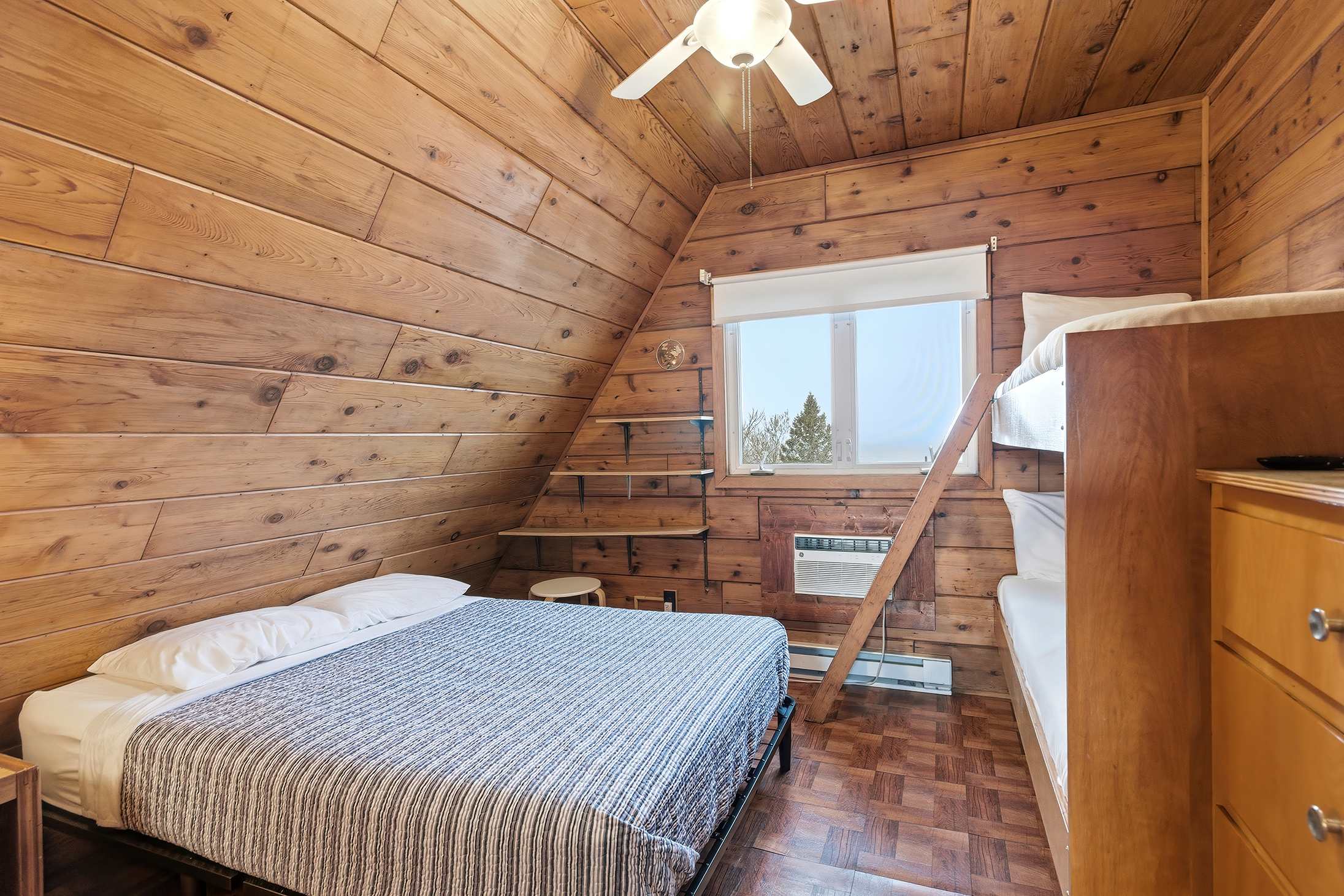 Bedroom-1 features rustic knotty pine walls and a sloped ceiling, furnished with a double bed and a wooden bunk bed. A window provides natural light, and a ceiling fan is mounted overhead.