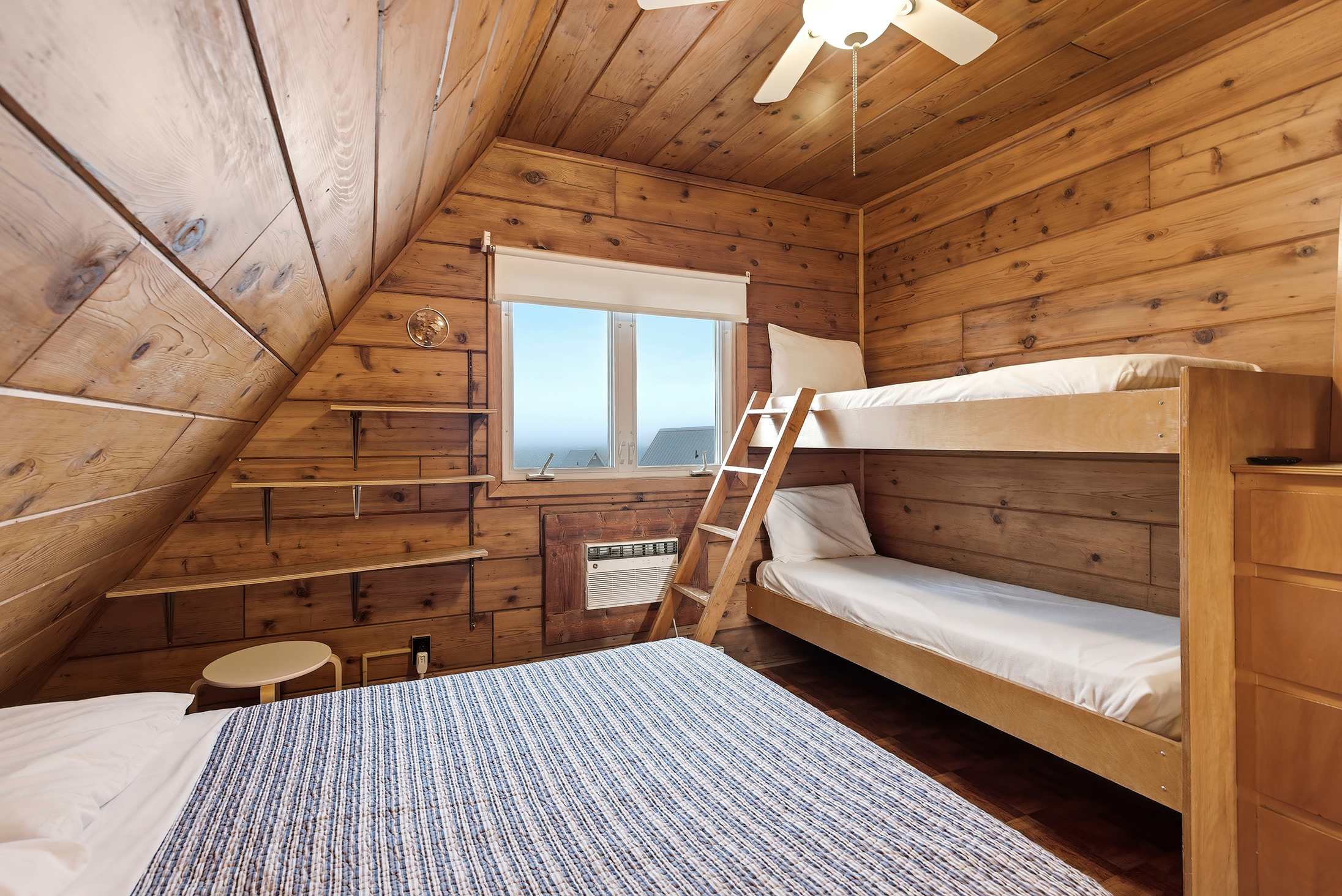 A cozy bedroom with sloped wood-paneled walls and ceiling, featuring a full bed with a blue and white striped comforter, and a wooden bunk bed. A window offers a view of the outdoors, and an AC unit is visible below it.