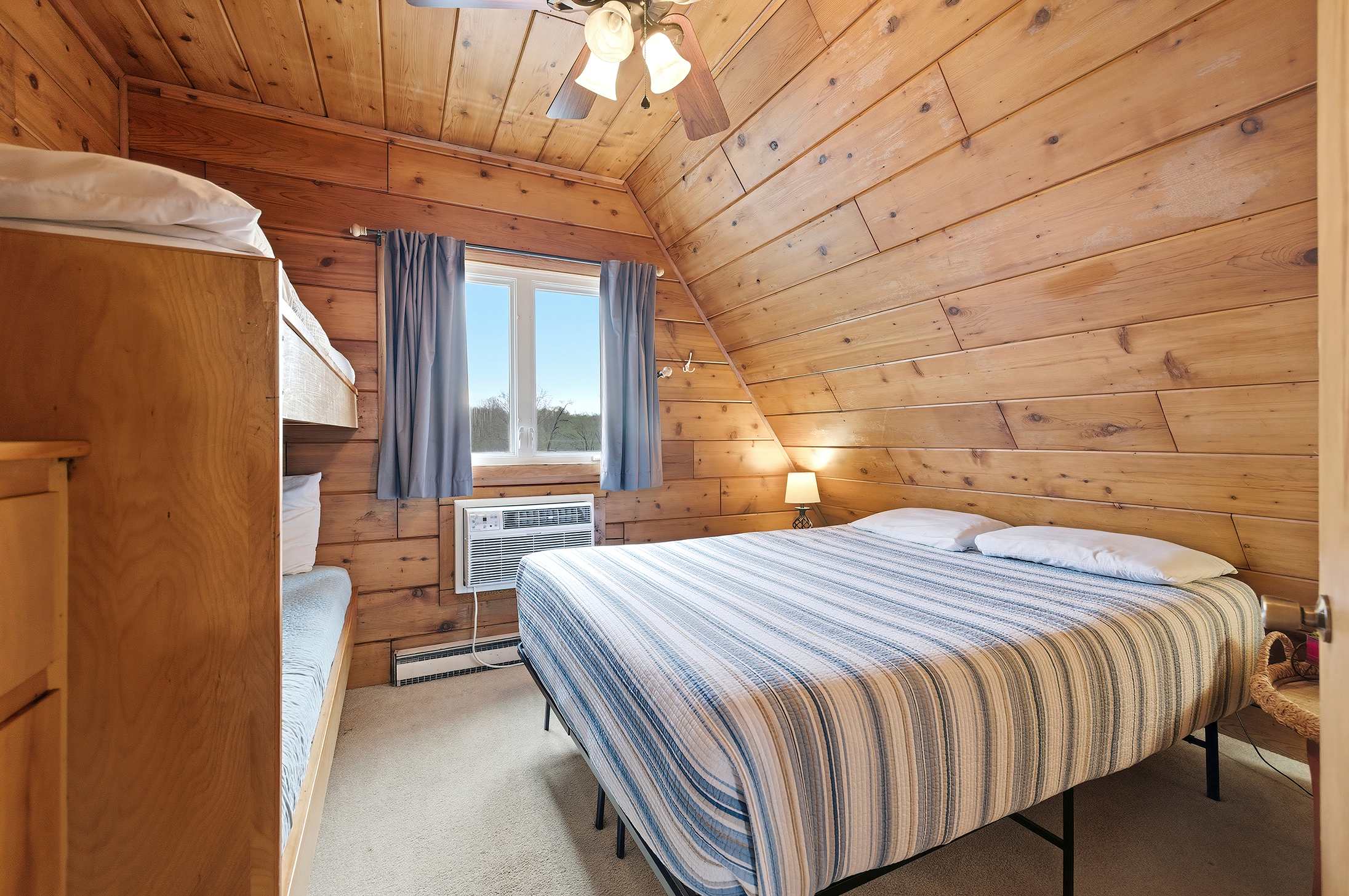 Cozy bedroom with warm wood paneling on walls and a vaulted ceiling, featuring a queen bed with blue and white striped bedding, a window AC unit, and a set of bunk beds on the left.