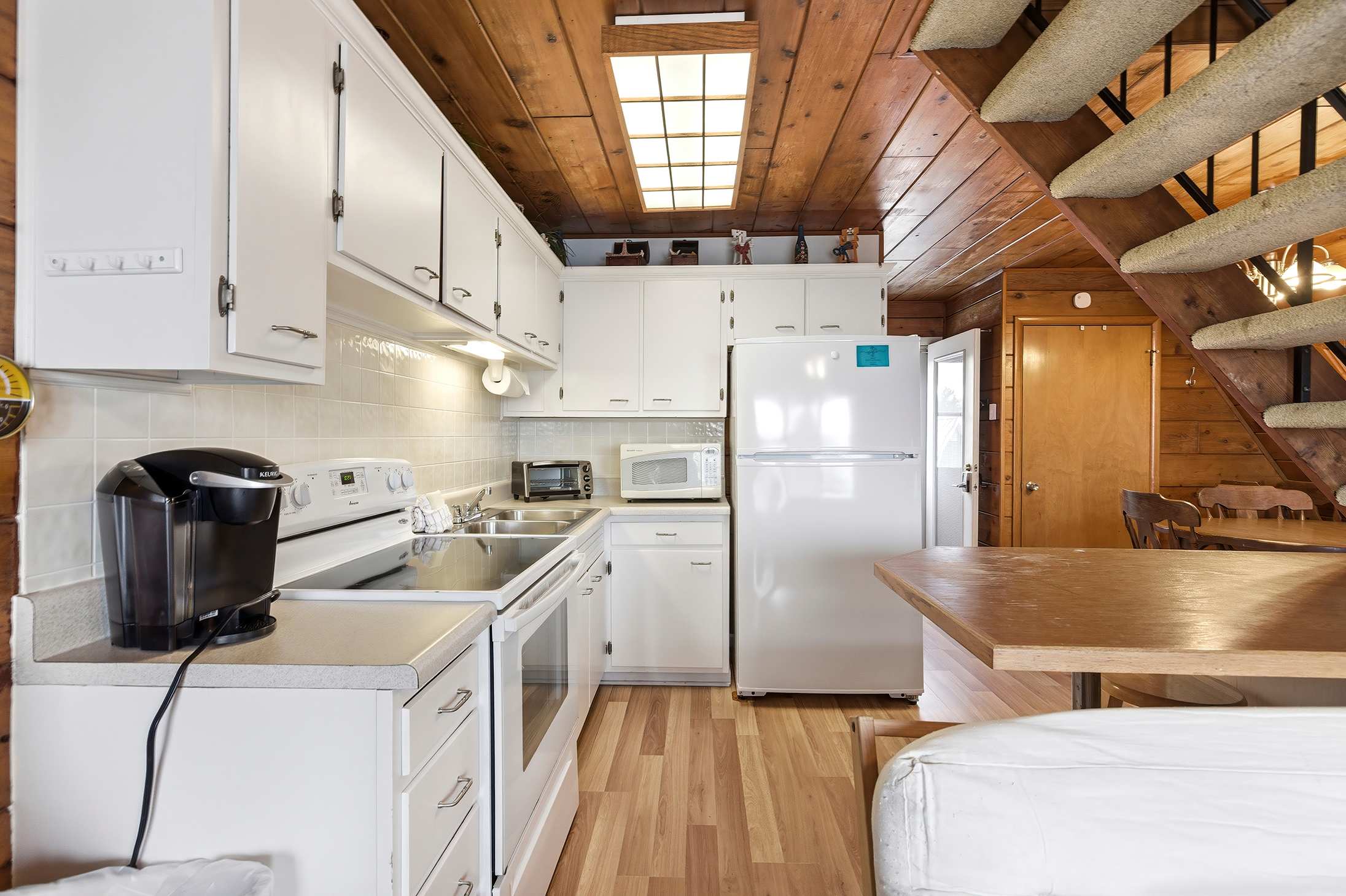 Bright kitchen area with white cabinets and appliances, including a refrigerator, stove, and microwave, set against wood-paneled walls and ceiling. A carpeted staircase with a black railing leads to an upper level, and a small dining counter is visible.