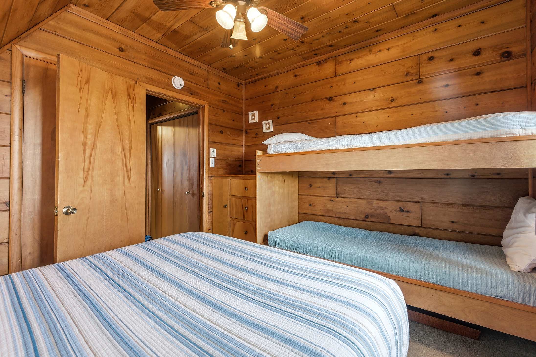 Alternate view of the wood-paneled bedroom, showcasing a queen bed with striped bedding, a wooden bunk bed with blue mattresses, and a built-in wooden closet and drawers.
