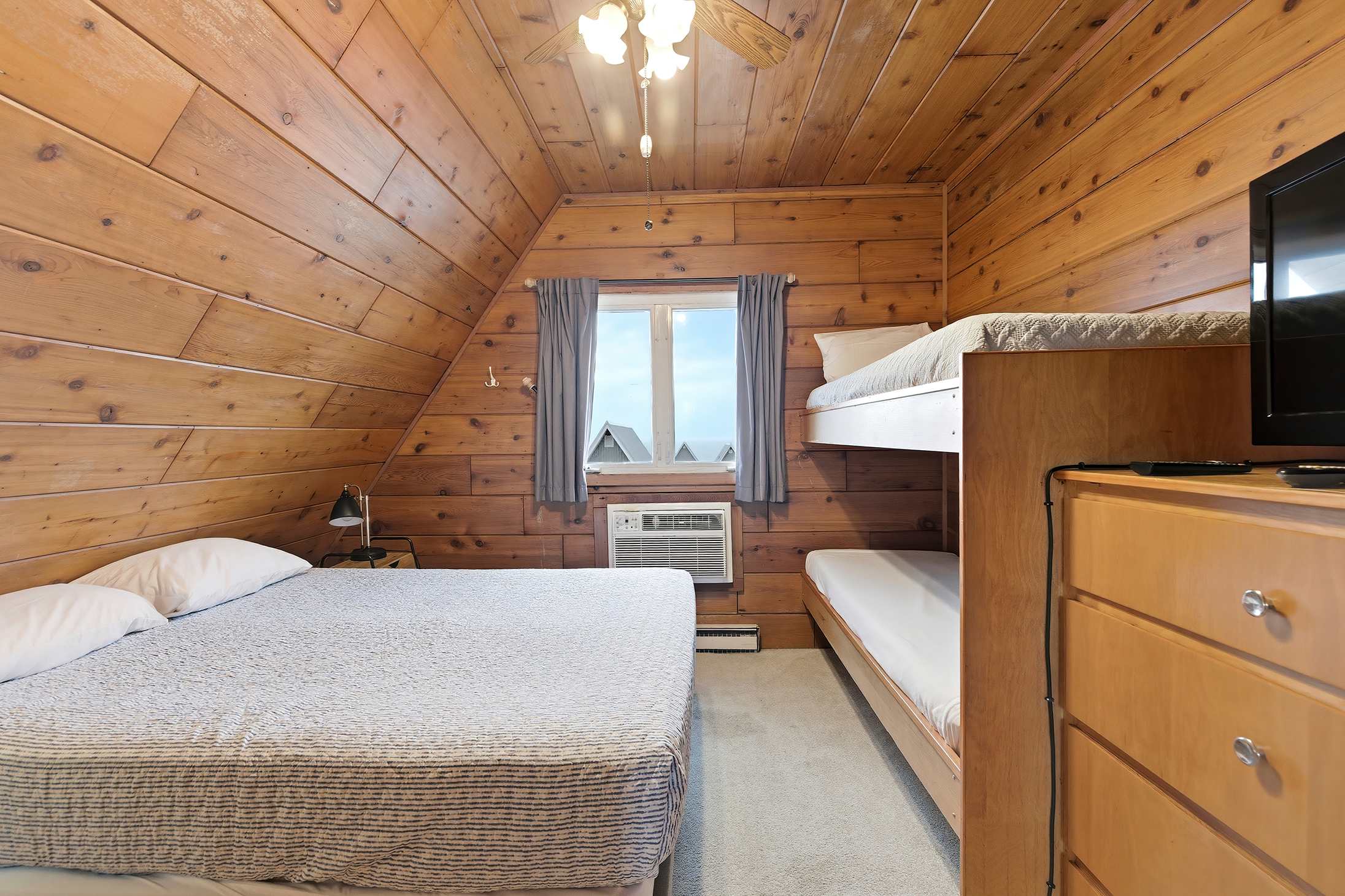 Rustic bedroom with warm wood-paneled walls and a sloped ceiling, furnished with a queen bed and a wooden bunk bed. A window with blue curtains provides a view, and a small TV is placed on a dresser next to the bunk beds.