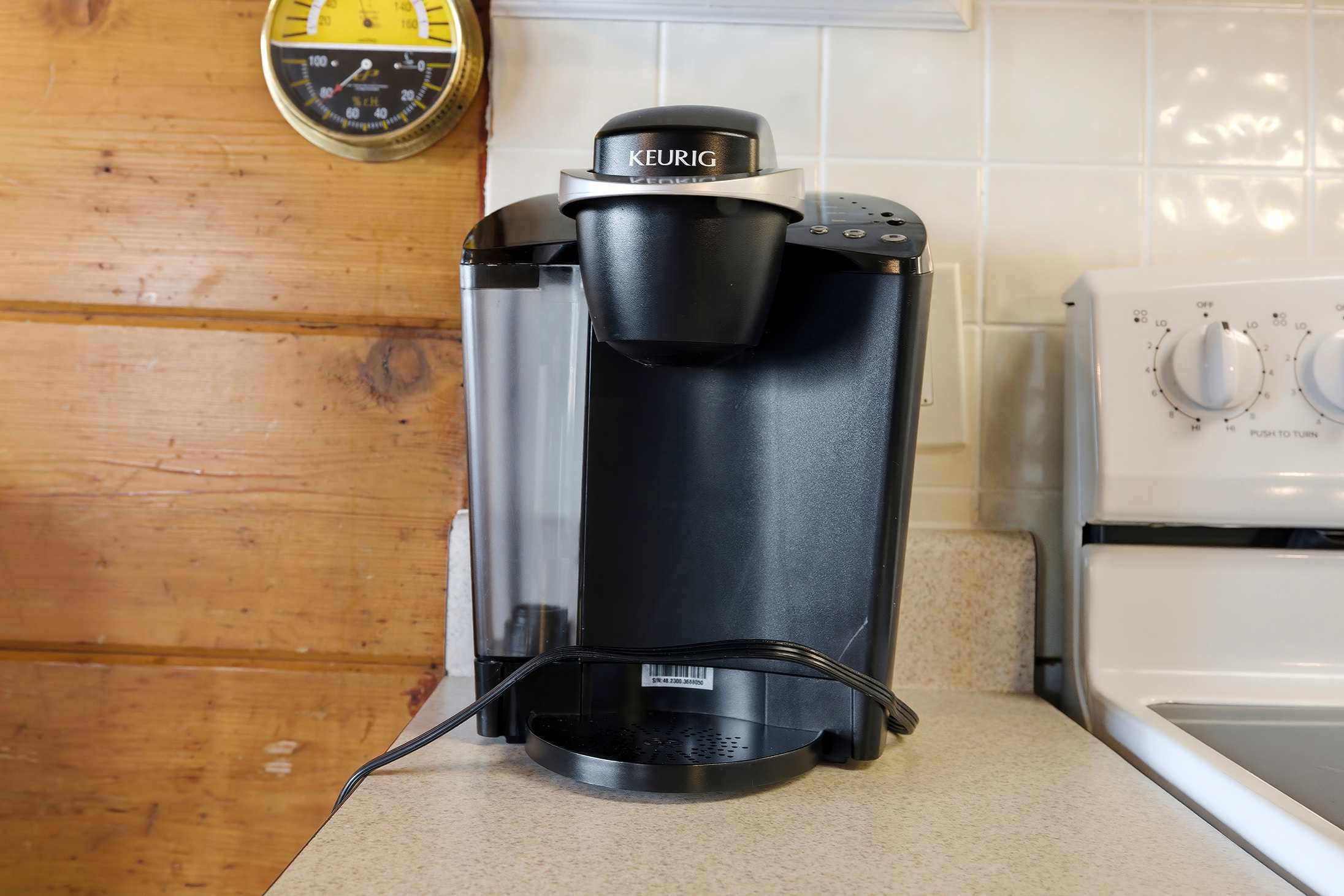 Close-up view of a black Keurig coffee maker sitting on a light-colored countertop in the kitchen, with a white electric stove and a yellow wall thermometer visible in the background. The wall is clad in warm wood paneling.