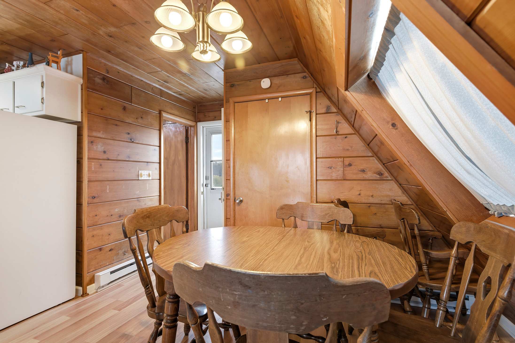 Rustic dining area featuring a round wooden table and chairs, surrounded by warm wood-paneled walls and a sloped wood ceiling. A classic chandelier hangs above the table, and a closed wooden door is visible in the background.
