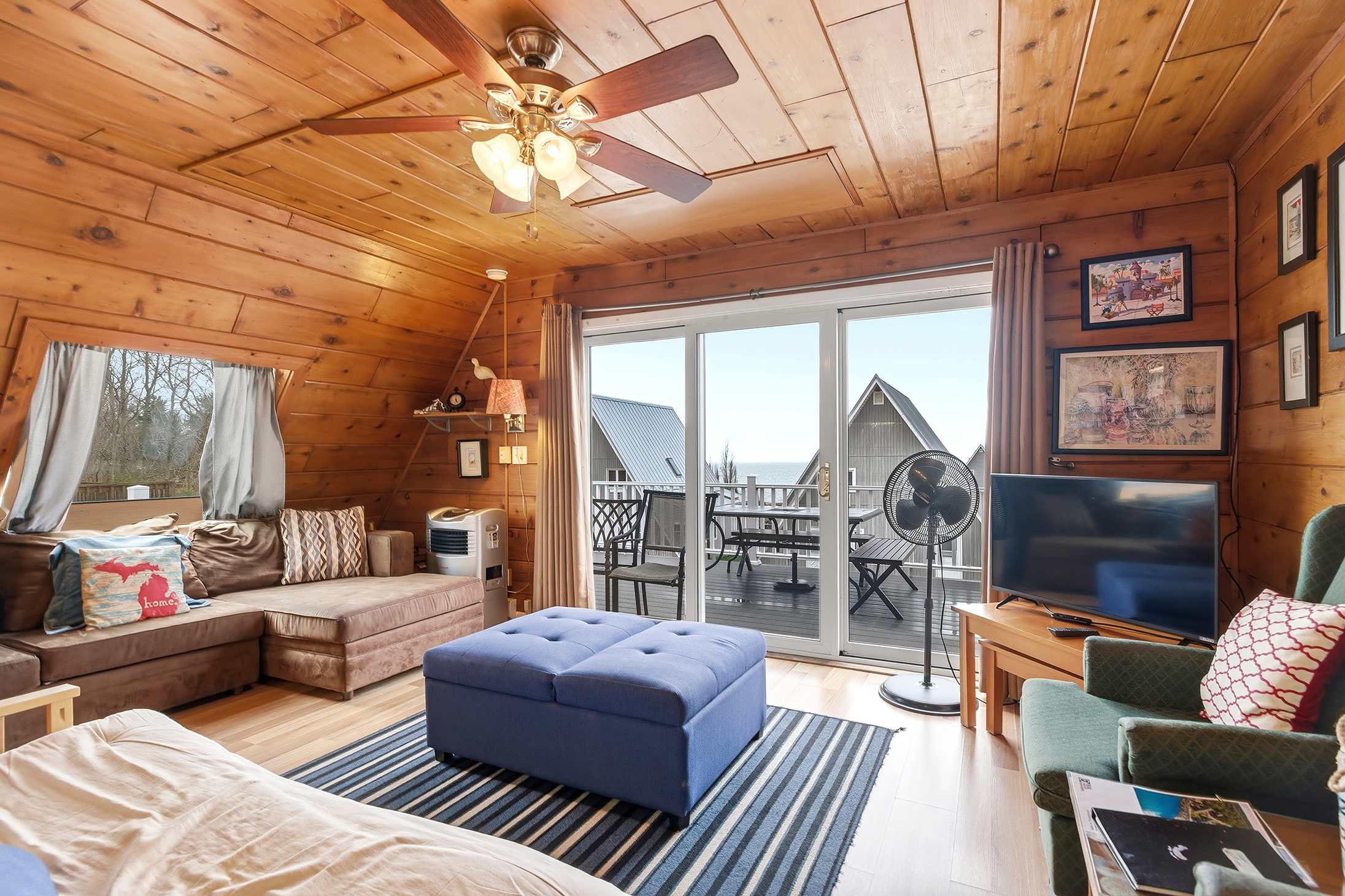 Inviting living room with extensive wood paneling on walls and ceiling, featuring a large L-shaped sectional sofa, a blue storage ottoman, and a sliding glass door opening to a deck with outdoor seating and a view of other chalets. A ceiling fan and floor fan are present.