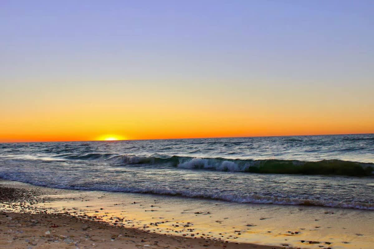 Scenic view of a sunset over a large body of water, with orange and yellow hues on the horizon, gentle waves breaking on a sandy and pebbly shore, and a clear sky fading to blue above.