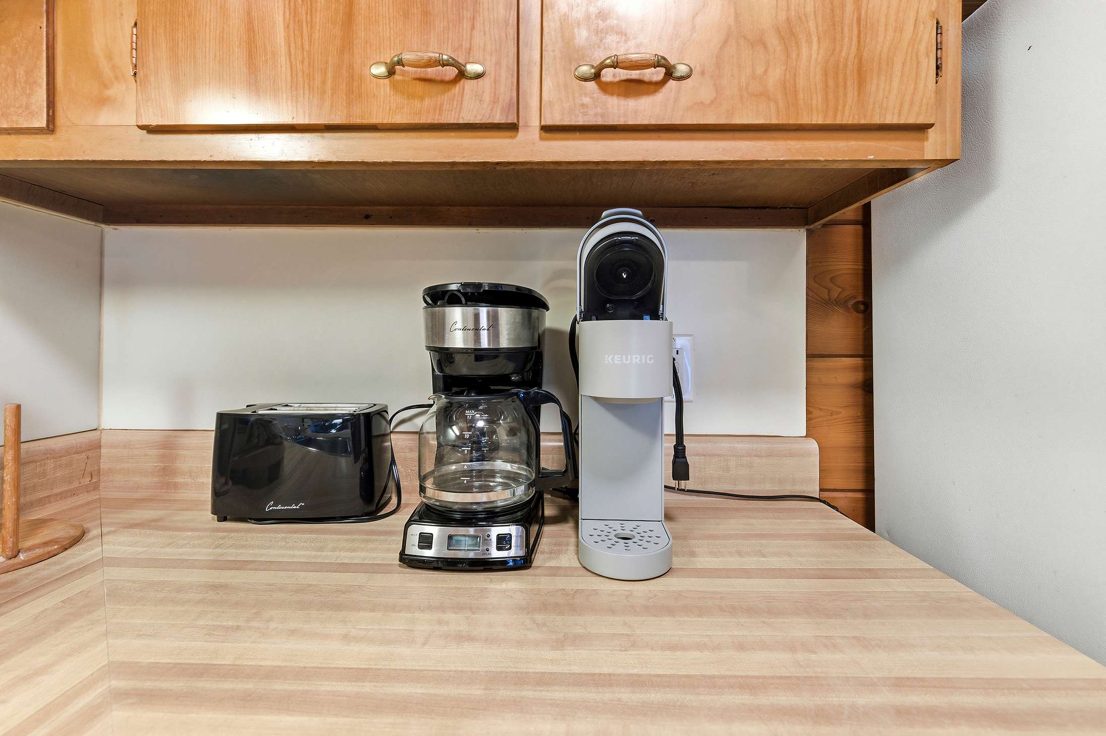 A functional kitchen counter features light wooden upper cabinets, a black toaster, a drip coffee maker, and a gray Keurig machine, all resting on a light wood-grain laminate countertop.