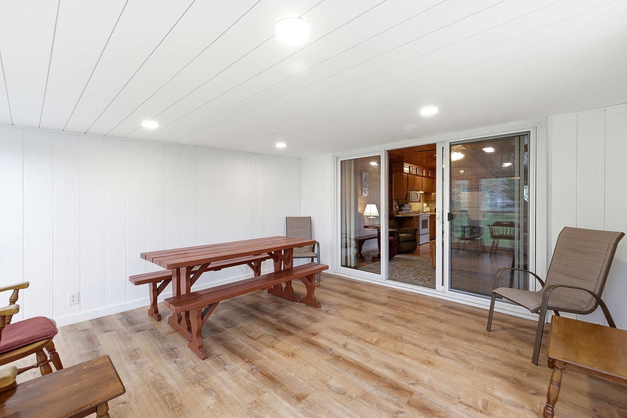 Enclosed porch with white paneled walls and light wood-look flooring, featuring a rustic wooden picnic table and benches, and a sliding glass door connecting to the main living area.