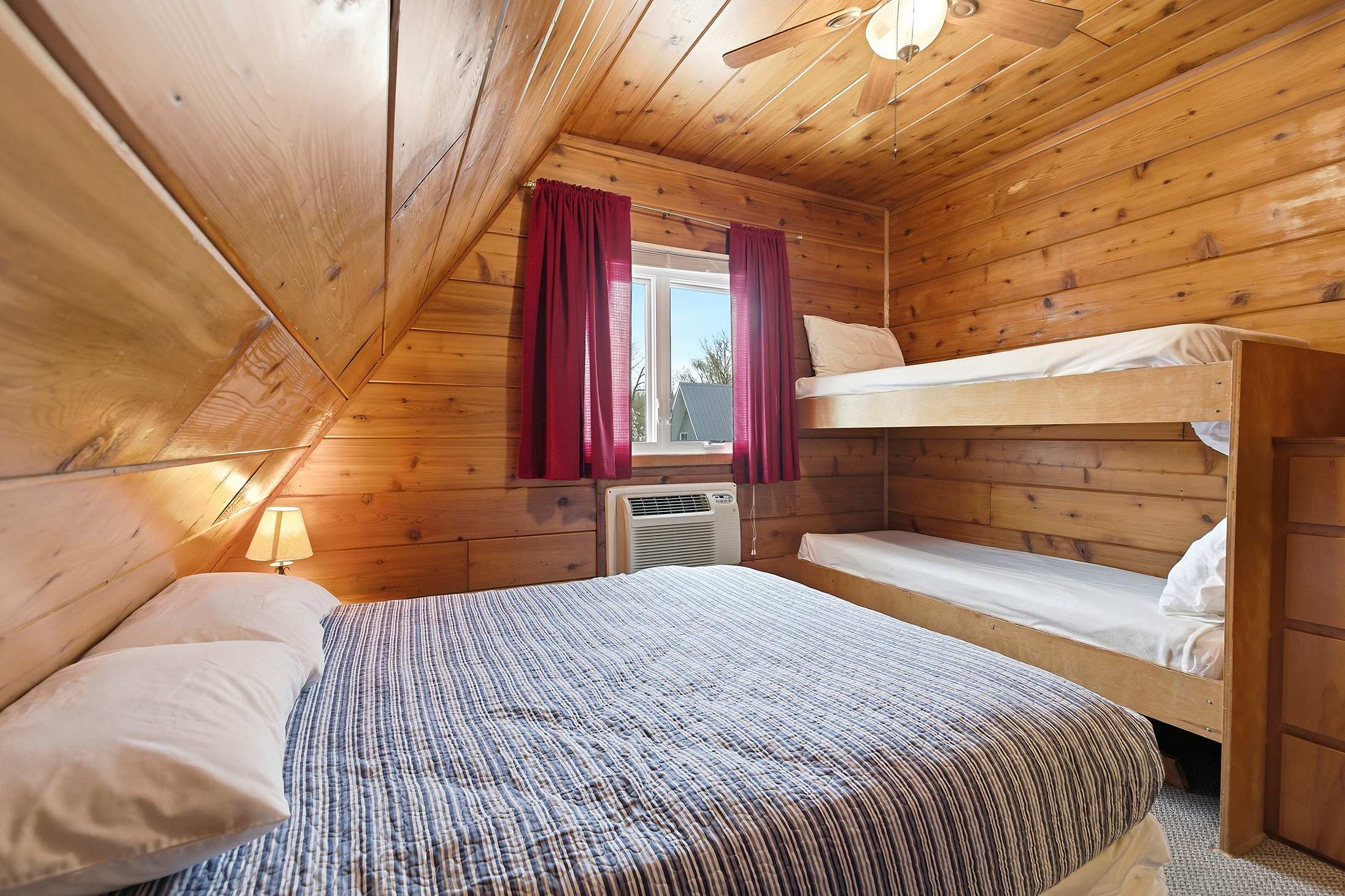 This cozy, wood-paneled bedroom features a double bed with a blue and white striped comforter, a wooden bunk bed, and a window with red curtains and a window-mounted AC unit, all beneath a sloped wooden ceiling.