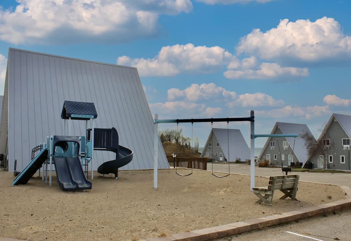 Modern playground with blue and gray slides and swings on a sandy base, featuring a wooden bench and several A-frame chalets visible in the background under a partly cloudy sky.