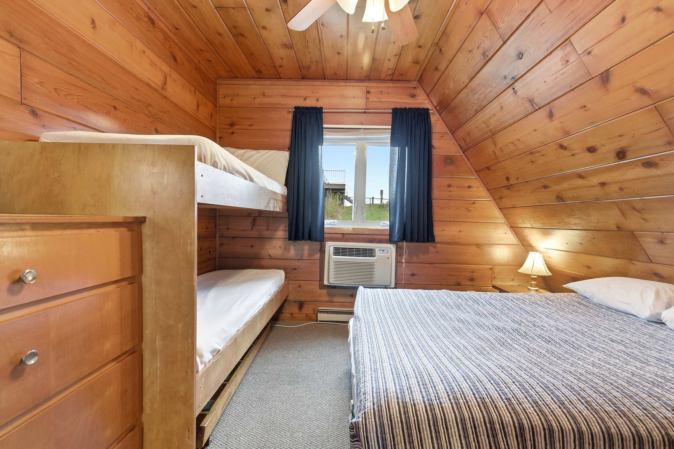 A rustic bedroom with warm wood-paneled walls and a sloped ceiling features a sturdy wooden bunk bed, a double bed with a blue and white striped comforter, and a window with blue curtains and a window-mounted AC unit.