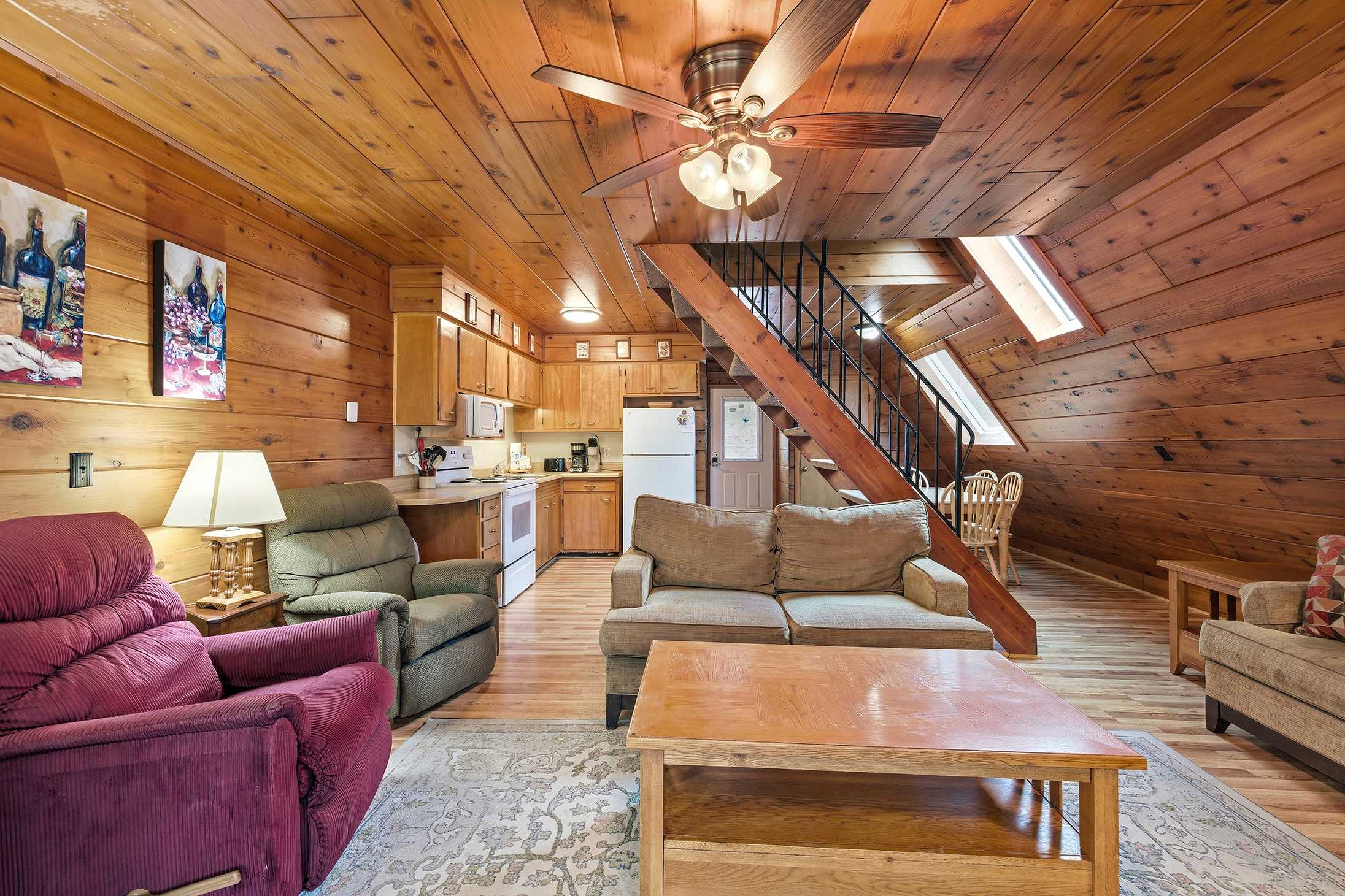 Open-concept living area and kitchen with extensive wood paneling, featuring a light brown sofa, a red recliner, a wooden coffee table, and a staircase with metal railing leading to an upper level.