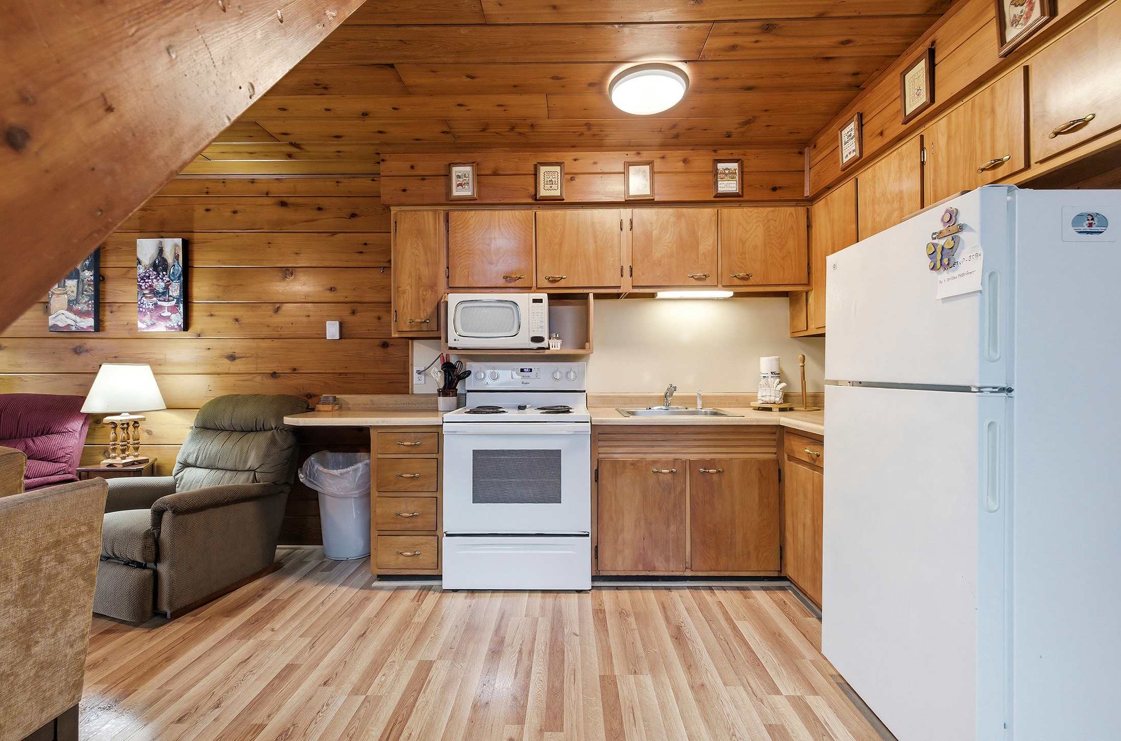 Functional kitchen area with light wood cabinetry, a white stove, microwave, and refrigerator, set against warm wood-paneled walls and ceiling, with a small counter space and sink.