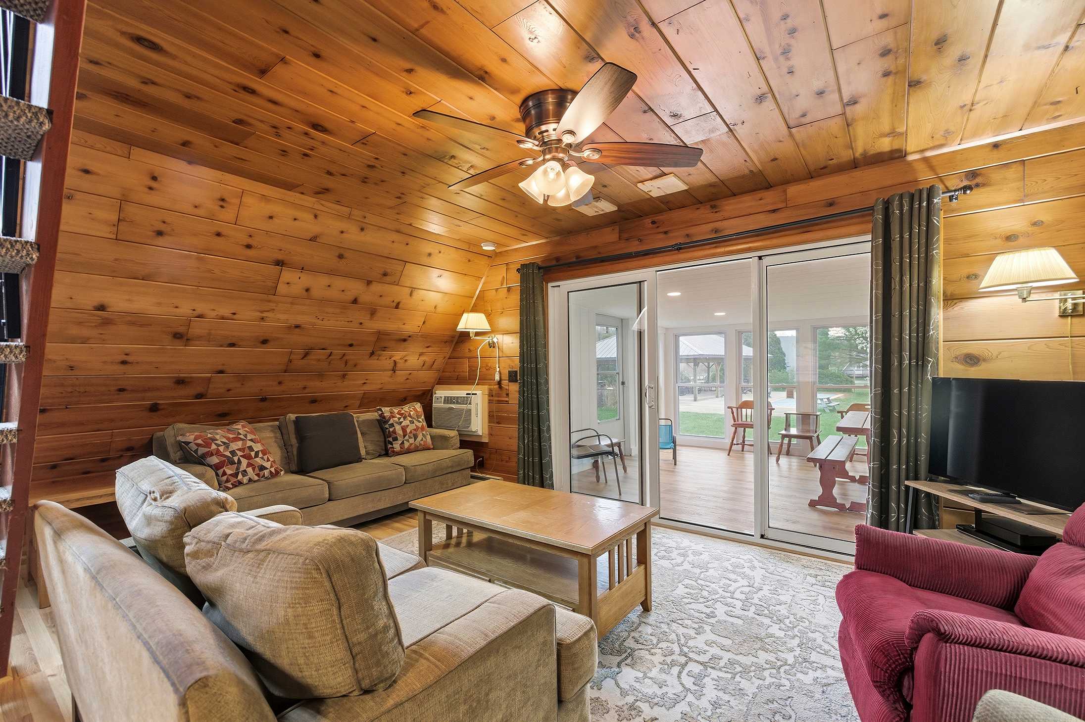 Inviting living room with warm wood-paneled walls and ceiling, furnished with a light brown sofa, a red recliner, a wooden coffee table, and a large sliding glass door opening to an enclosed porch.