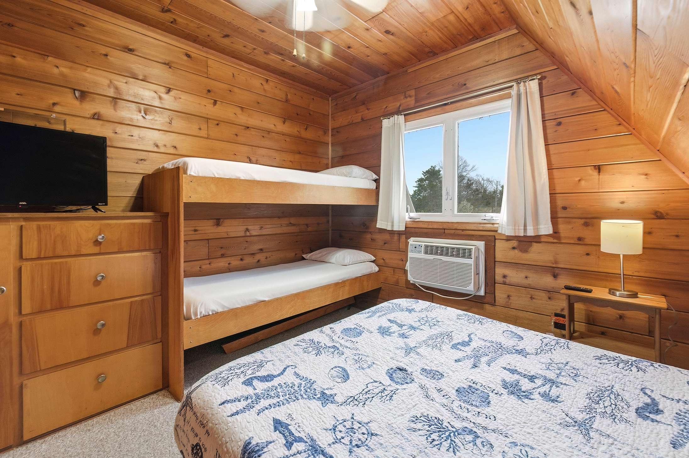 A cozy bedroom with knotty pine wood paneling on walls and ceiling, featuring a wooden bunk bed, a queen-sized bed with a blue and white patterned quilt, and a window AC unit. A wooden dresser with a TV sits to the left.