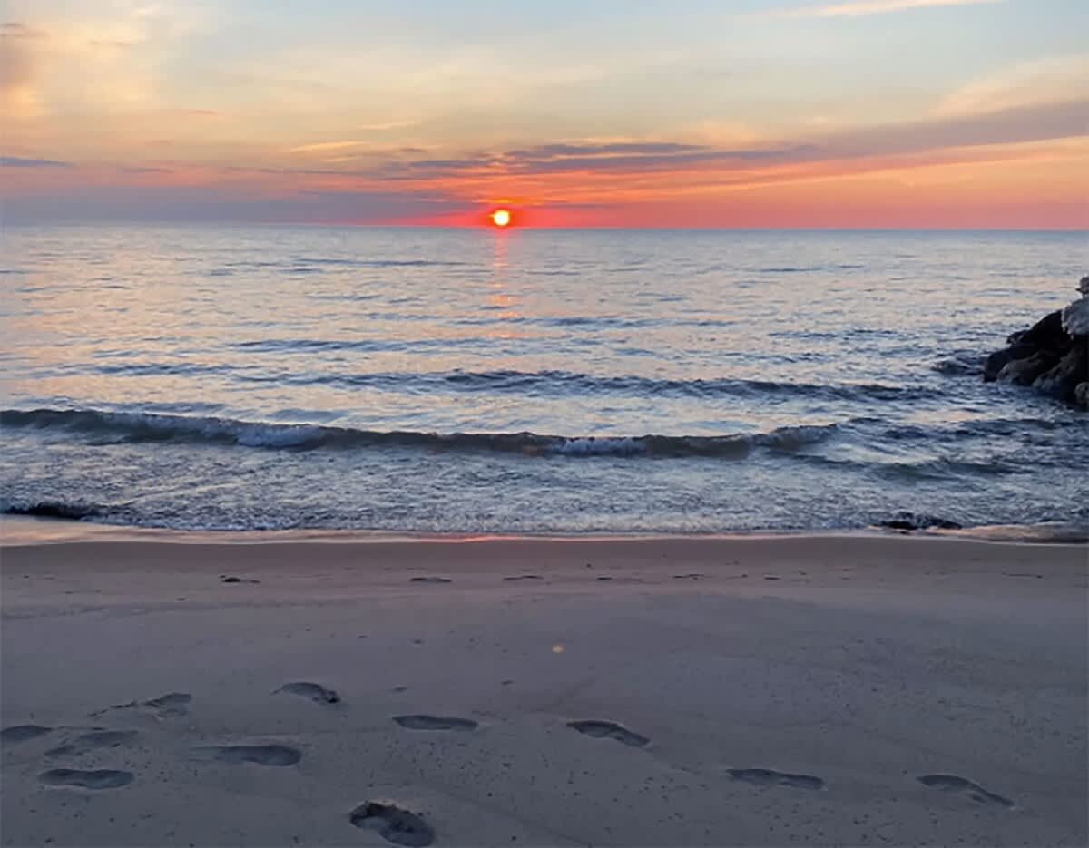 A serene sunset over a vast body of water, with the sun dipping below the horizon casting an orange glow. Gentle waves lap at a sandy beach with visible footprints in the foreground.
