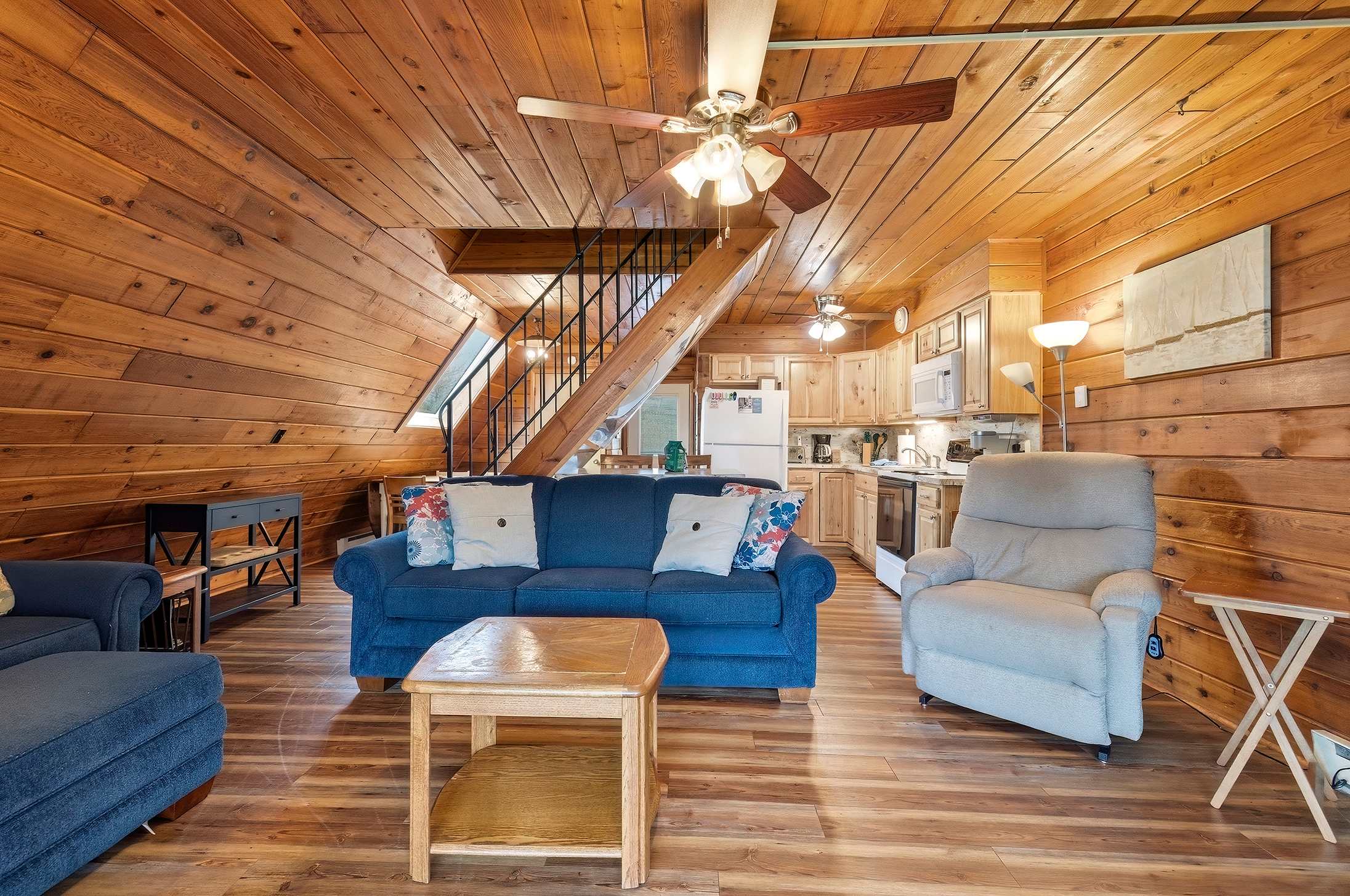 An open-concept living area boasts extensive wood paneling on walls and ceiling, a blue sofa, a recliner, and a wooden coffee table. A staircase with metal railings leads upstairs, and a compact kitchen is visible in the background.