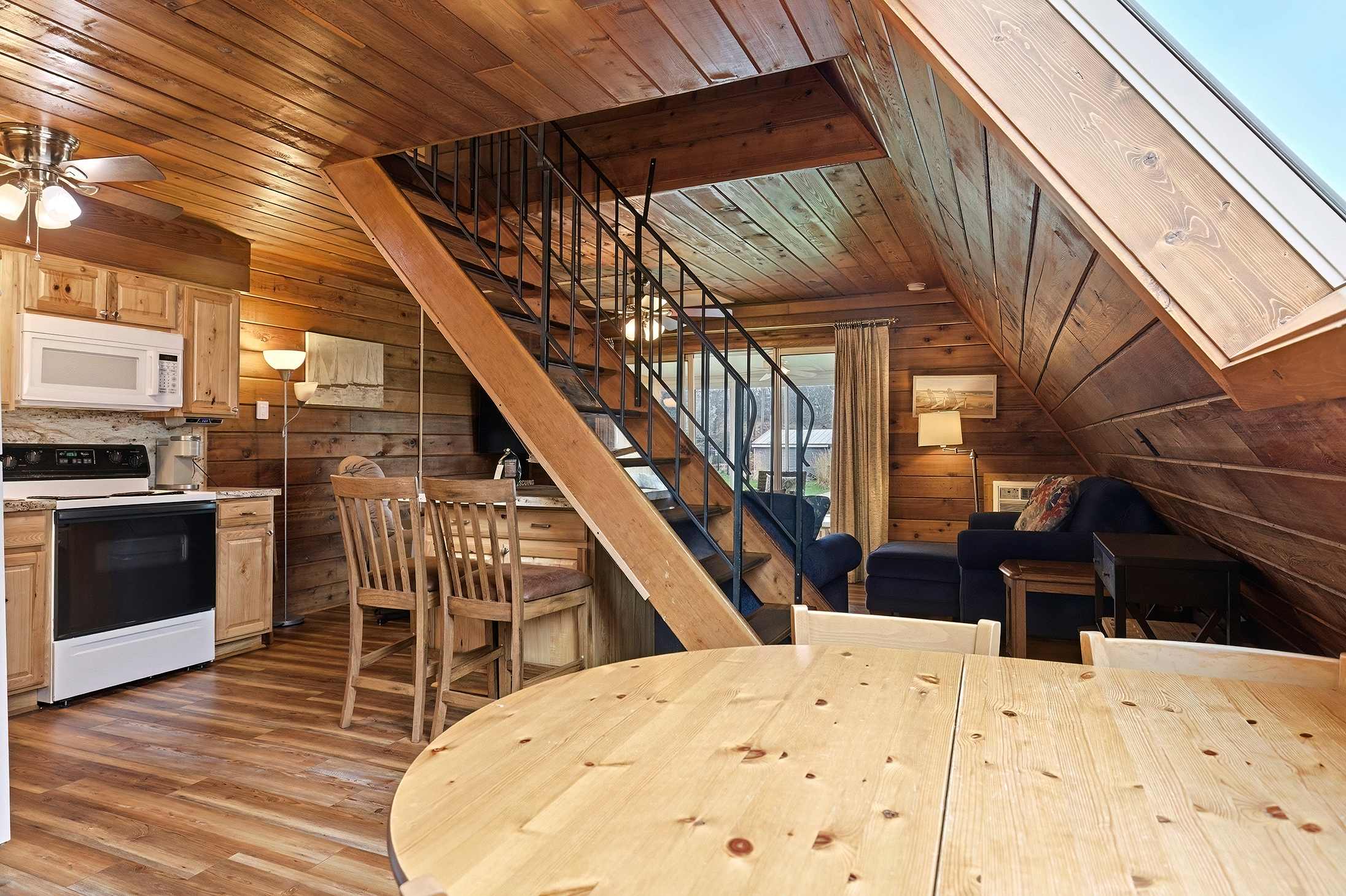 An open-concept living space showcases a dining area with a round wooden table, a compact kitchen with wood cabinetry, and a prominent staircase with black metal railings. The entire area is clad in warm wood paneling, creating a rustic cabin feel.