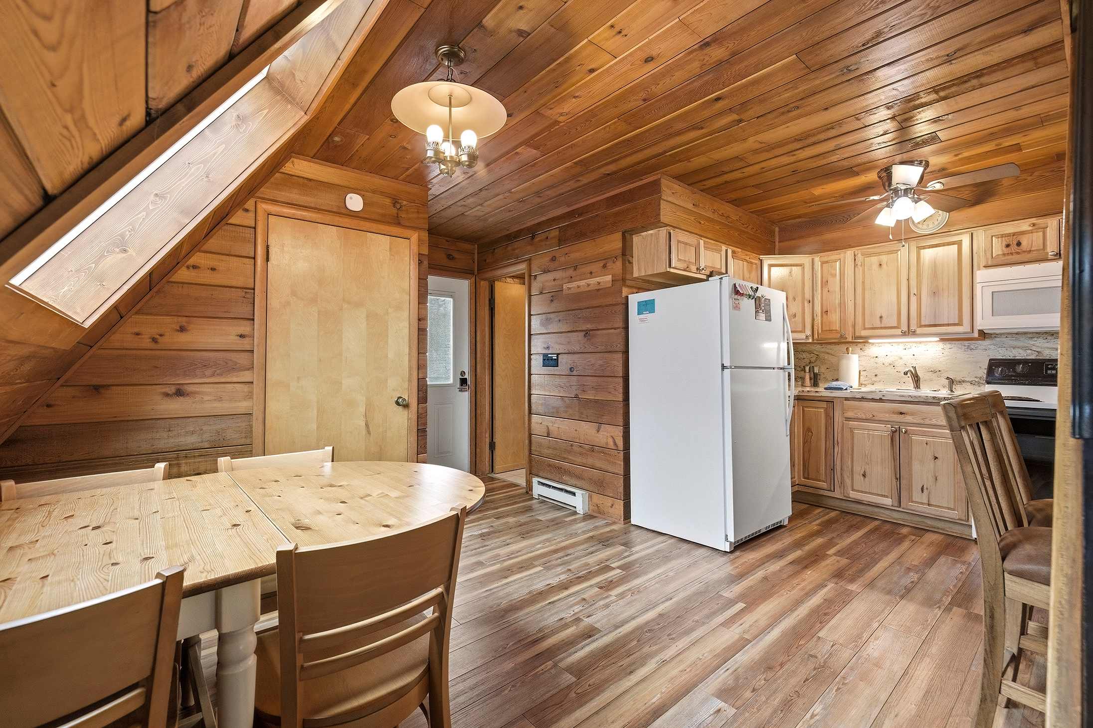 The dining area, with its light wood round table and chairs, flows into a compact kitchen featuring wood cabinets and a white refrigerator. The room is characterized by extensive wood paneling on walls and a sloped ceiling, with a door leading to another room.