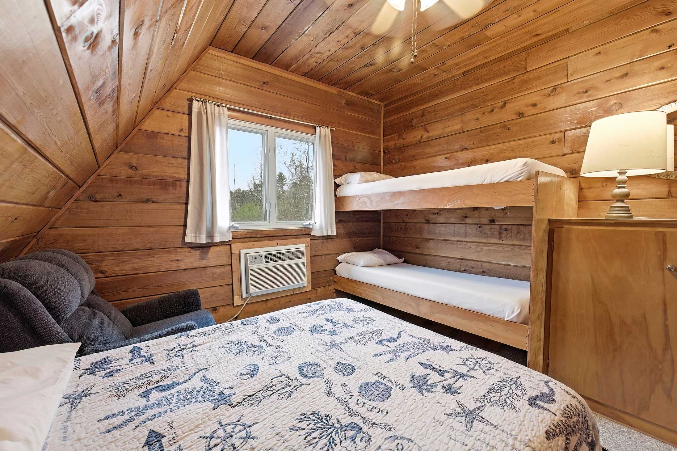View of a rustic bedroom with wood-paneled walls and ceiling, featuring a wooden bunk bed, a queen bed with a blue and white quilt, and a window with a window AC unit below. A dark gray armchair is visible in the corner.
